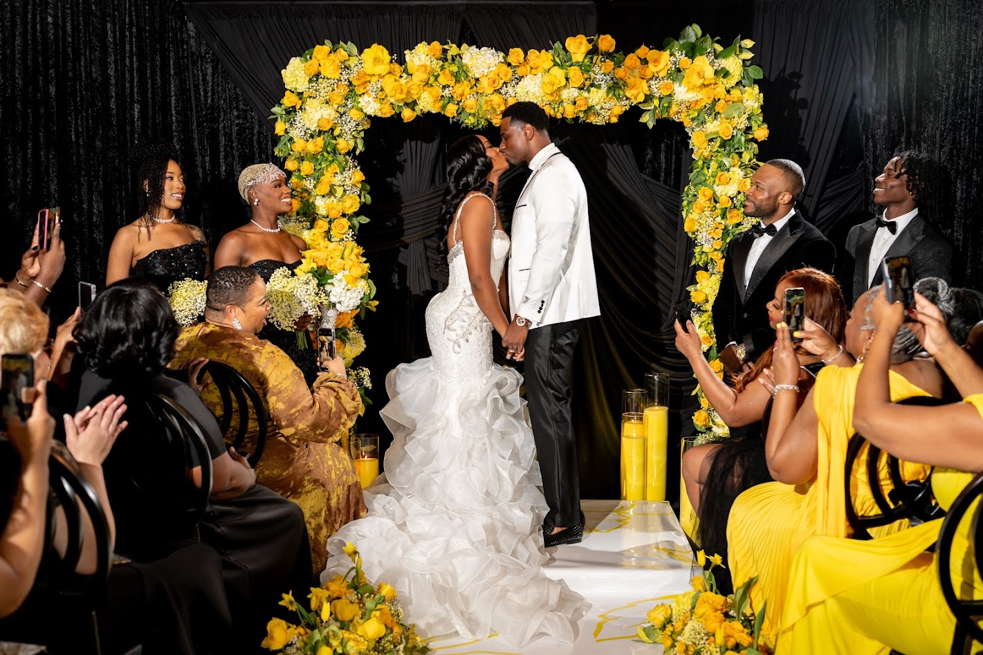 Bride and groom holding hands under a yellow floral arch during wedding ceremony, surrounded by guests clapping and taking photos.