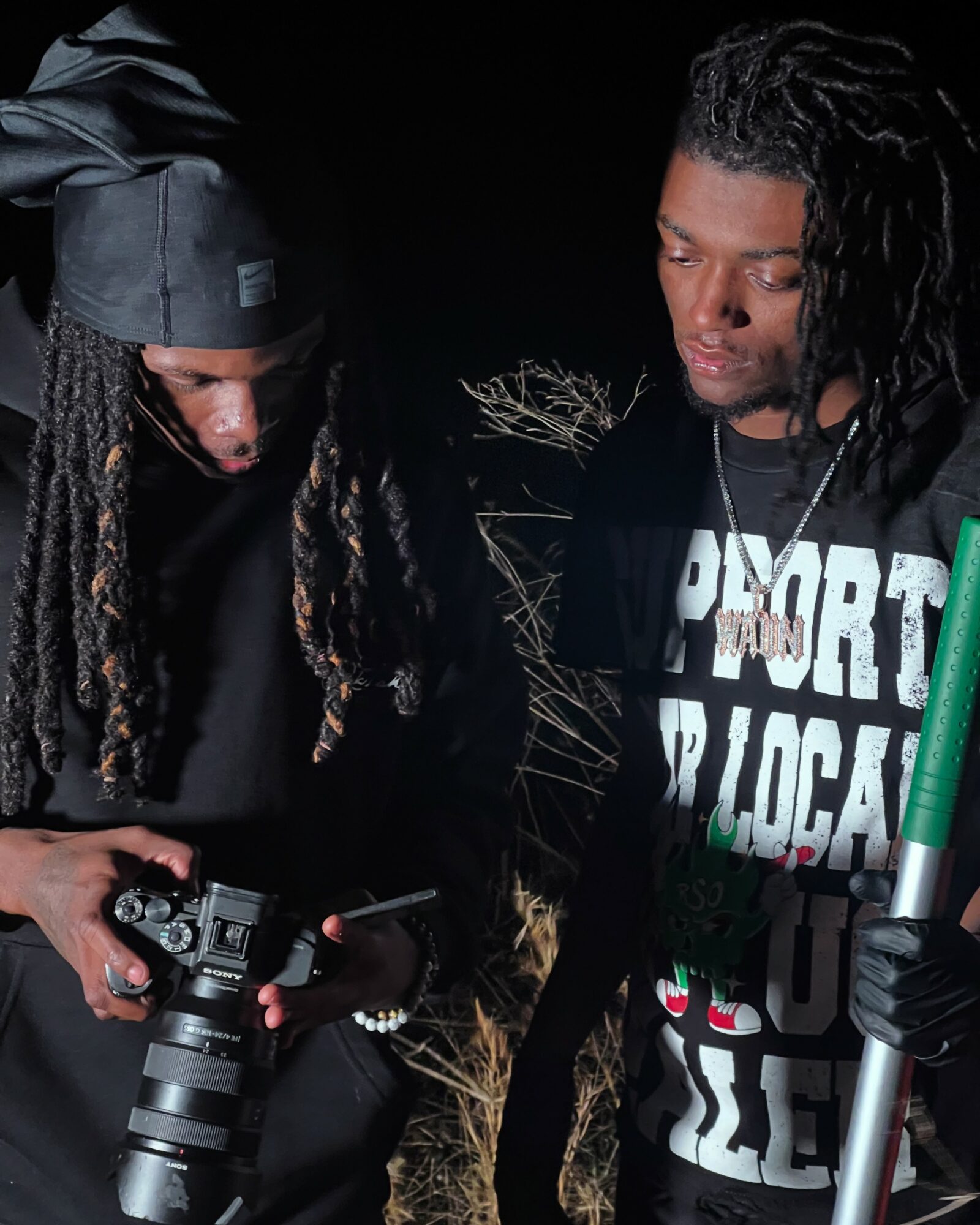 Two men with dreadlocks look at a camera and a phone against a dark background.