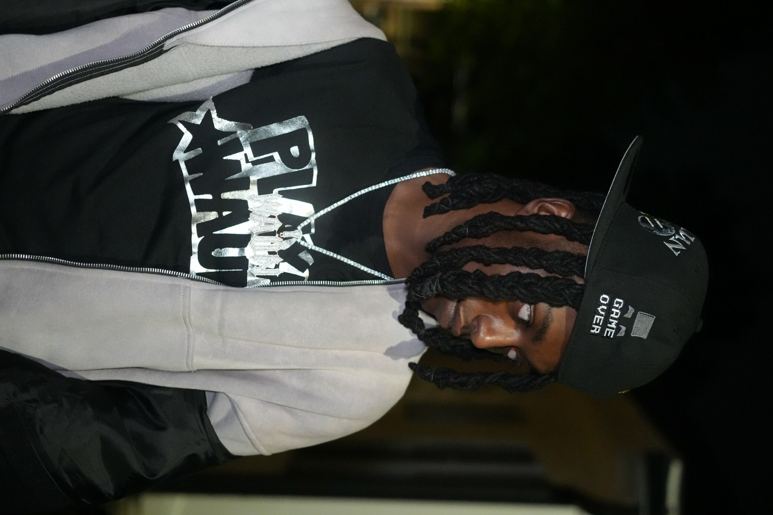 Person with braided hair wearing a cap, black T-shirt, and jacket, holding a phone outdoors at night.