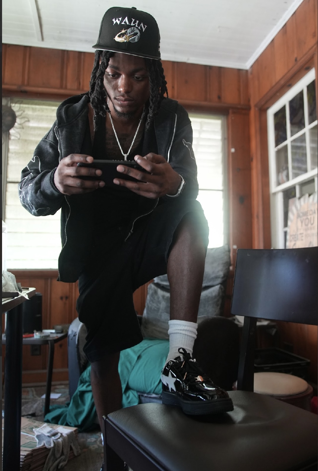 Person with dreadlocks wearing a cap, black jacket, and sneakers, standing on a chair indoors, looking at a phone.