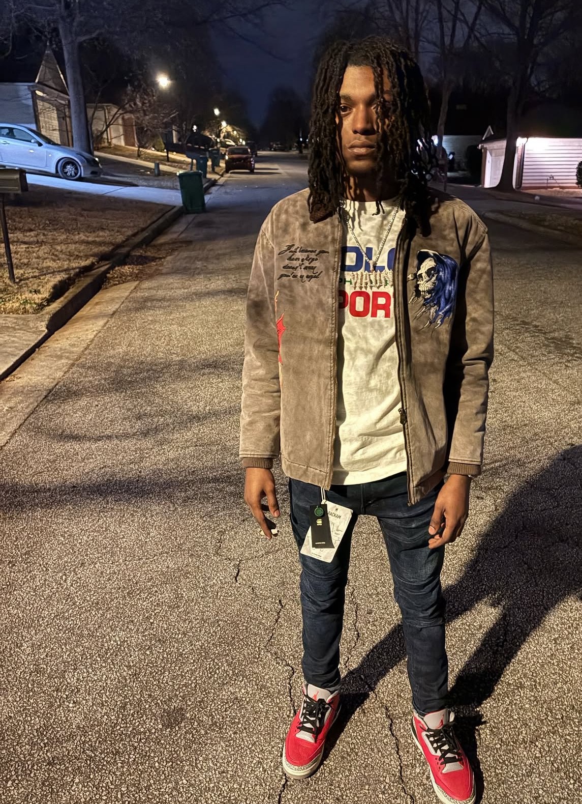 Young person with dreadlocks standing on a sidewalk at night, wearing a beige jacket, white shirt, dark pants, and red sneakers.