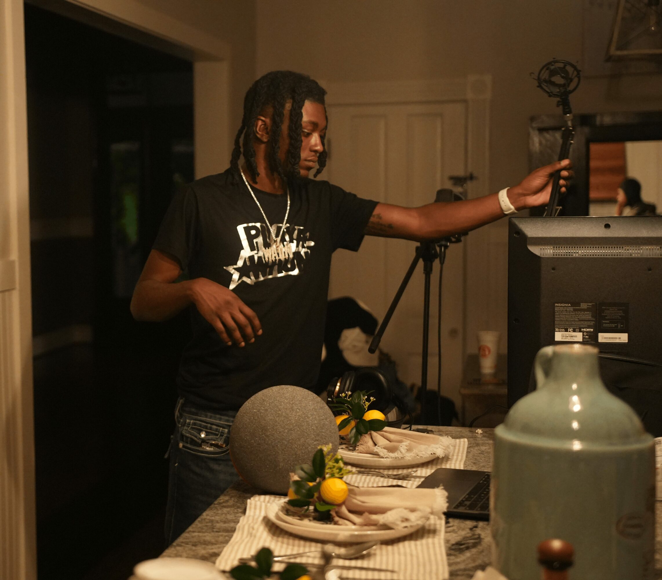 Person with dreadlocks wearing a black t-shirt, reaching towards a computer monitor in a dimly lit room.