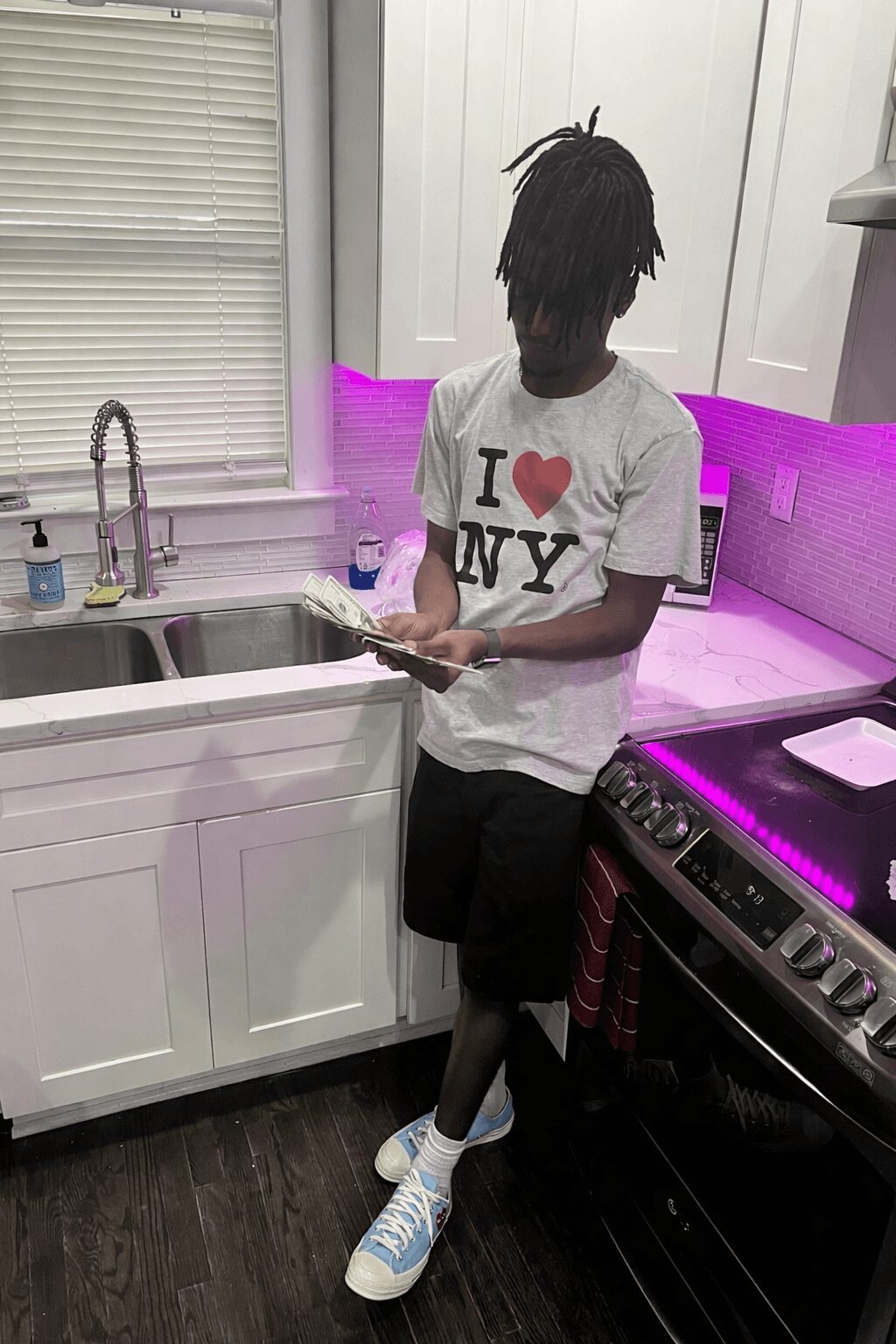 Young person with dreadlocks reading a book in a kitchen with white cabinets and purple lighting.