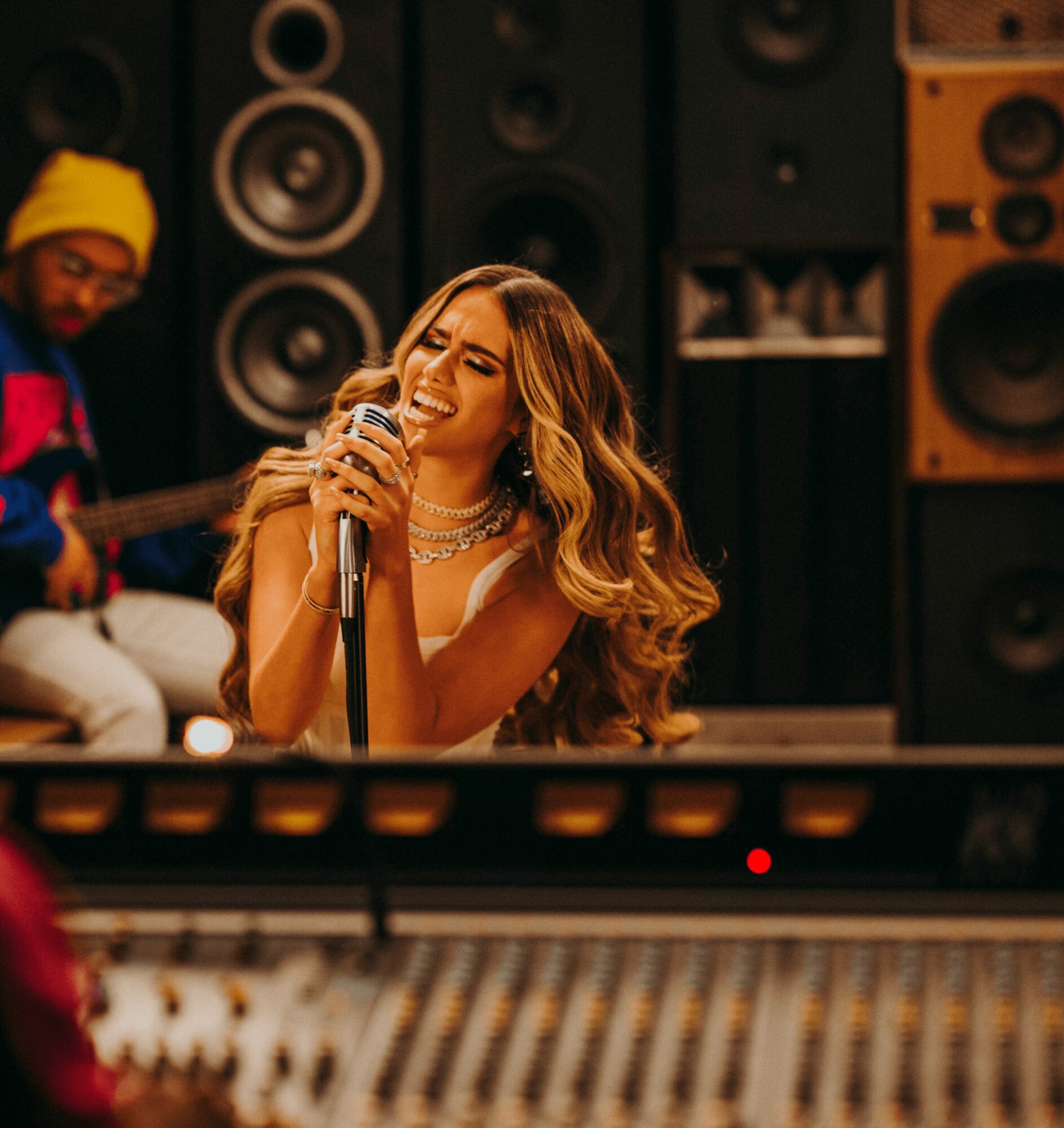 Woman singing into a microphone in a music studio with large speakers in background.