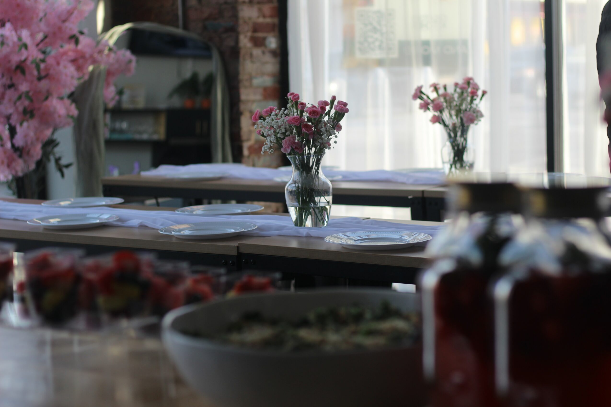 Tables with flower vases and papers, window with curtains in background, some food items in foreground.