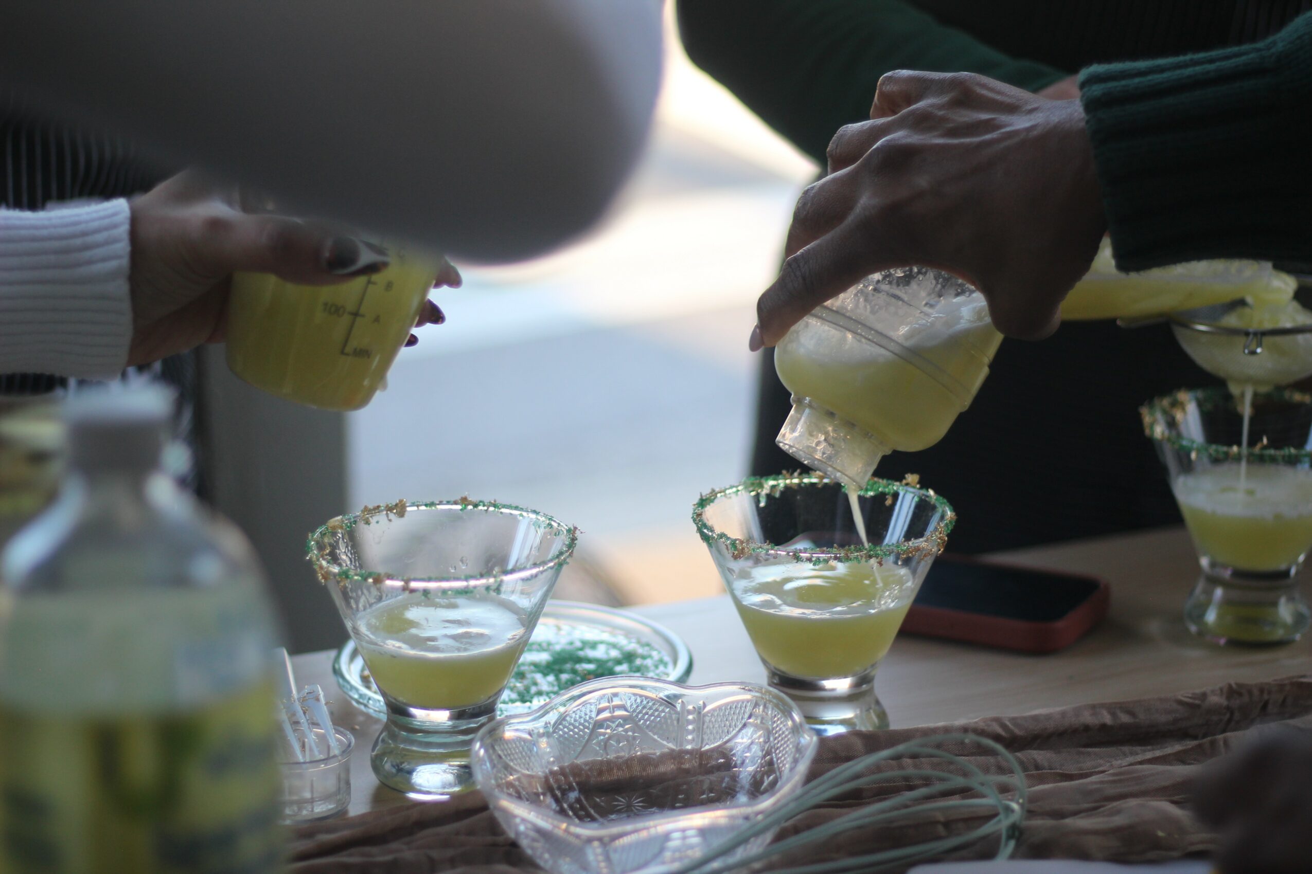 People pouring a yellowish drink into glasses with salted rims, with a blurred background and a bottle in the foreground.