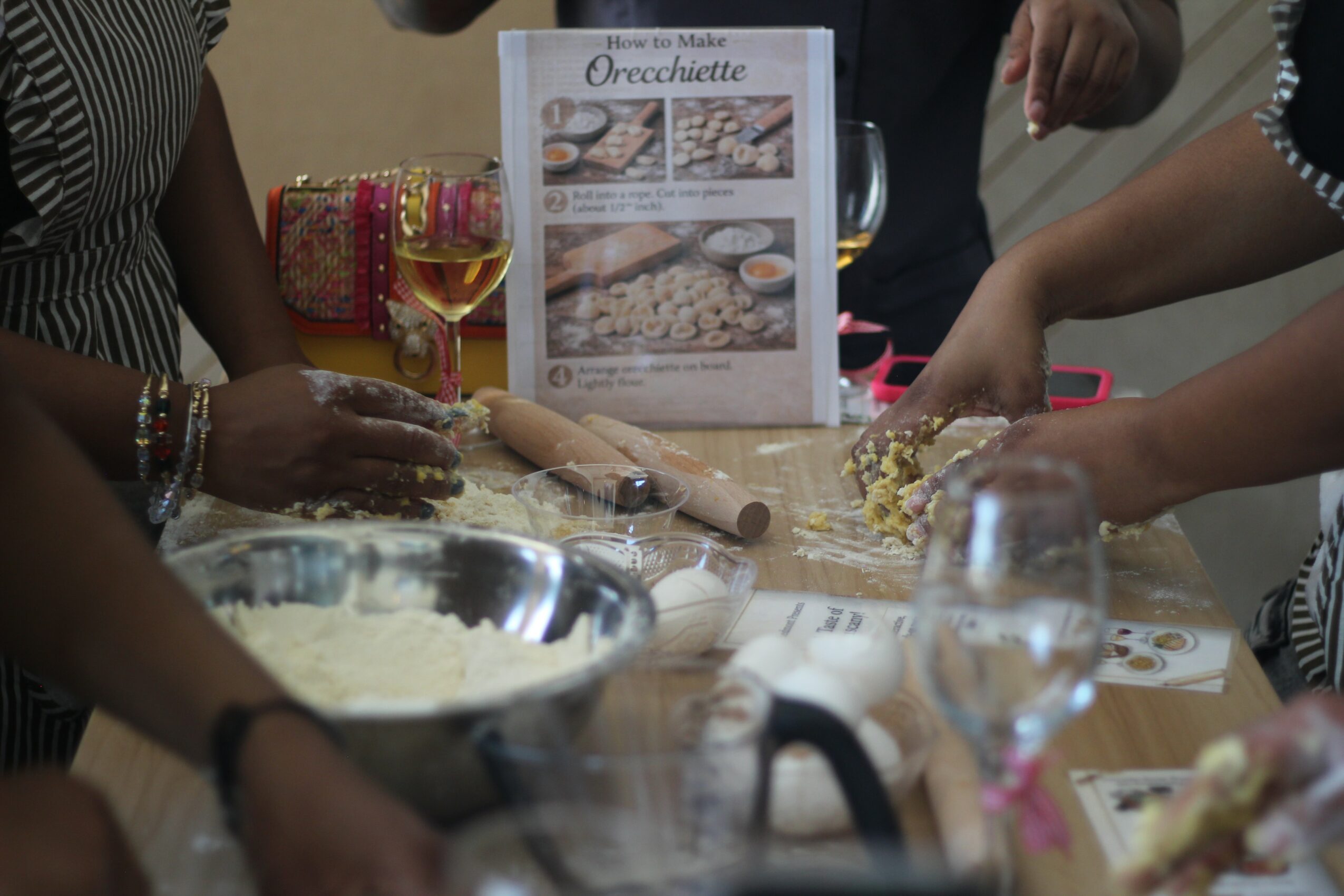 People gather around a table preparing to make orecchette pasta, with ingredients, utensils, and a menu displayed.