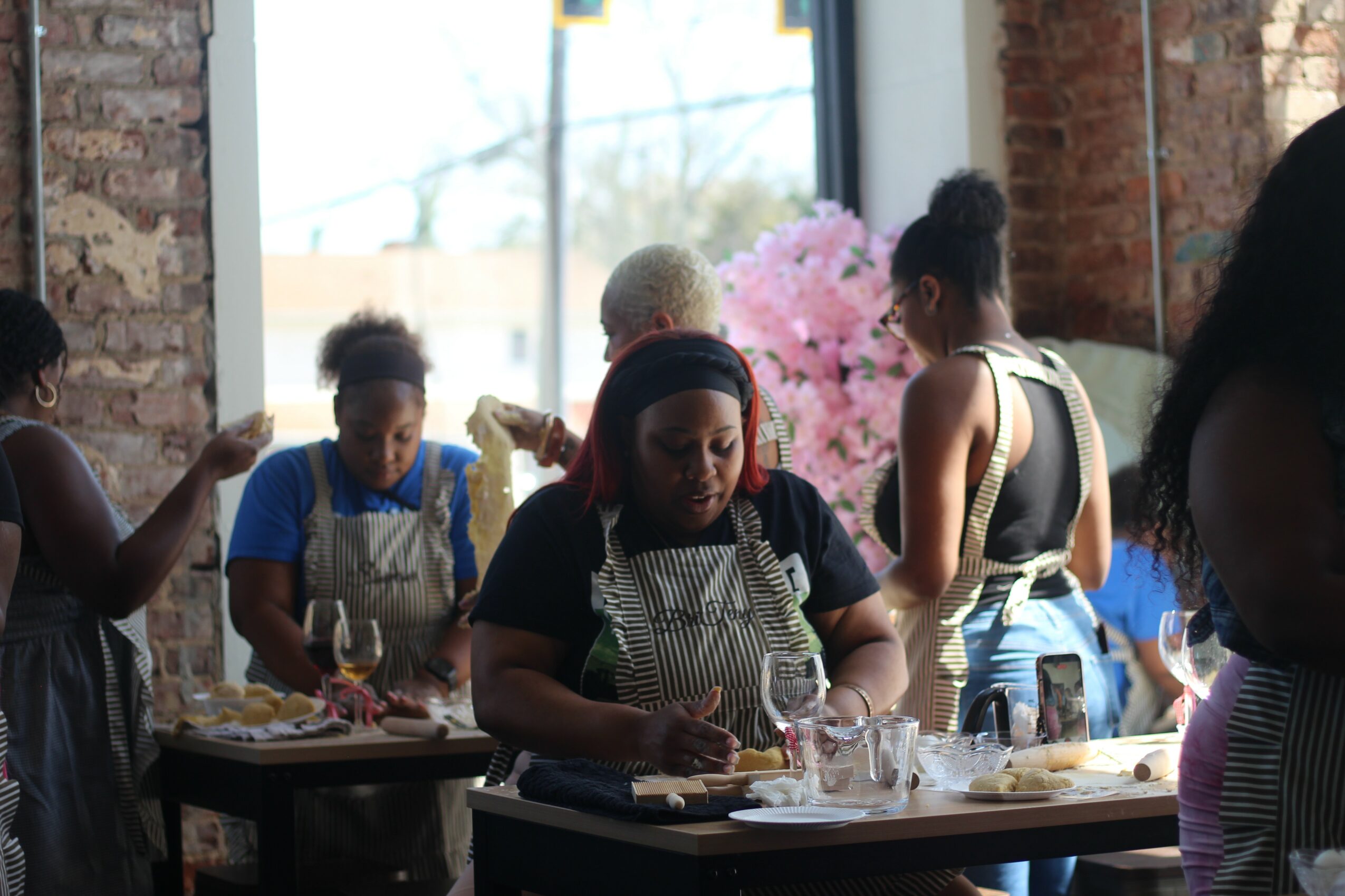 Group of women preparing food in a kitchen with brick walls and a window, pink flowers outside.