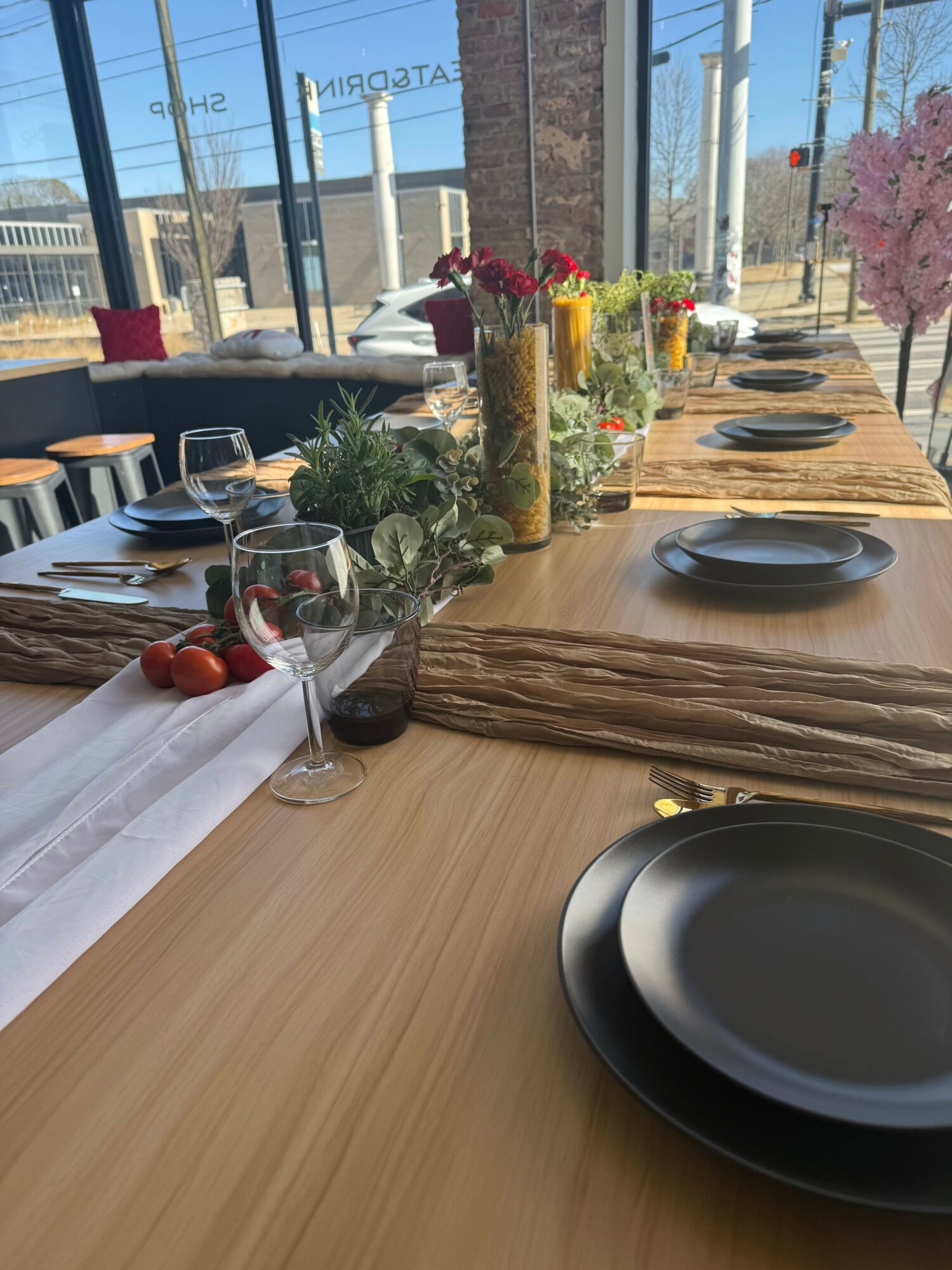 Long wooden table set with plates, glasses, and floral centerpieces in a bright room with large windows and pink blossoms outside.