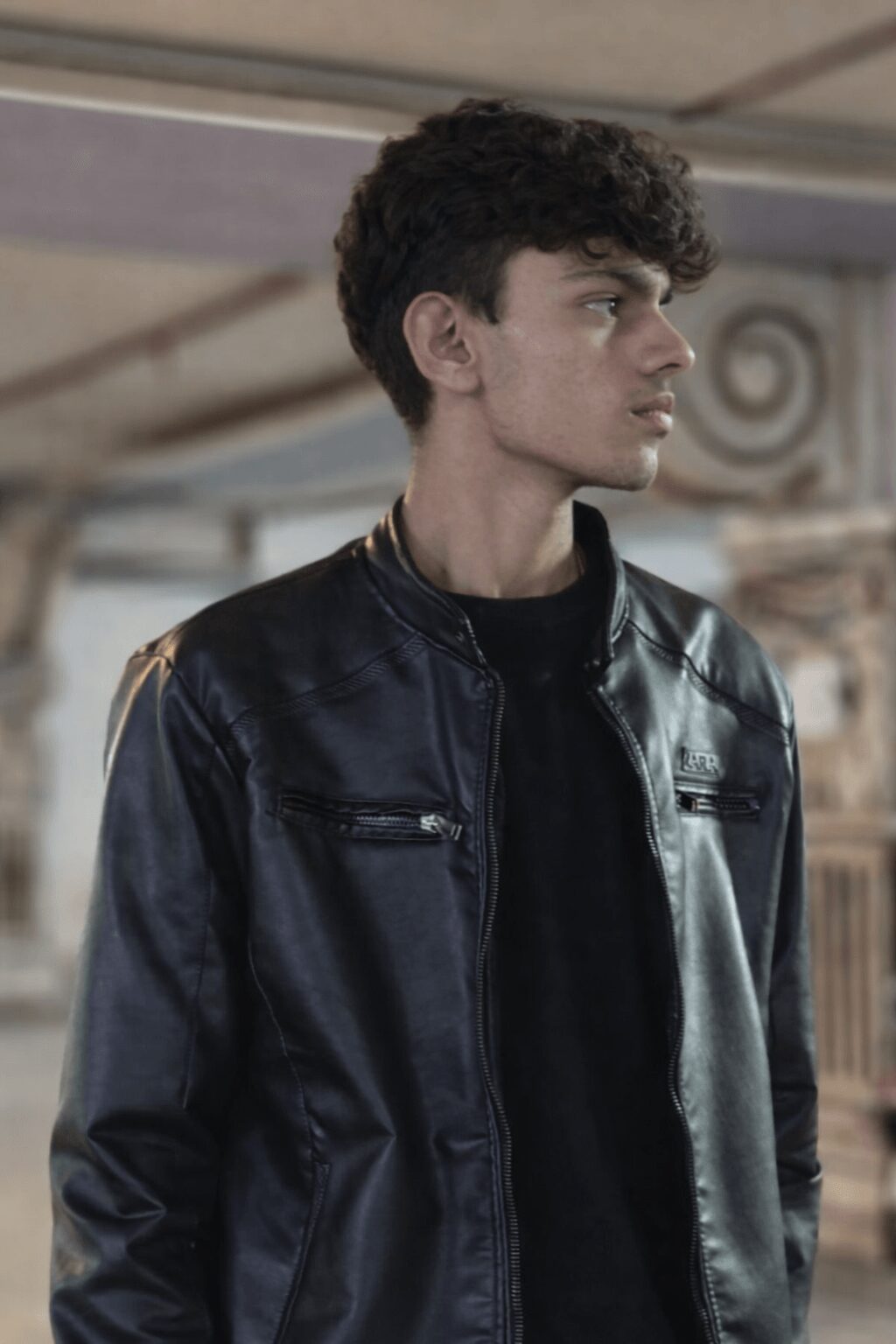 Young man with dark curly hair wearing a black leather jacket, looking to the right.