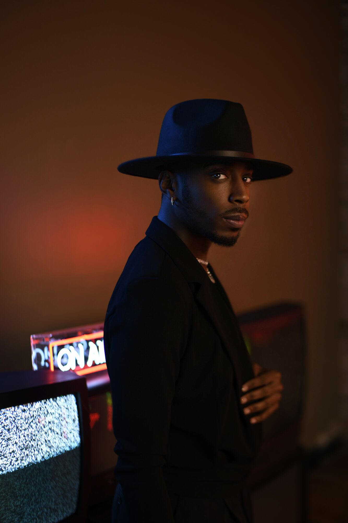 Person wearing a wide-brimmed hat and dark clothing, standing in a dimly lit room with a TV screen showing static.