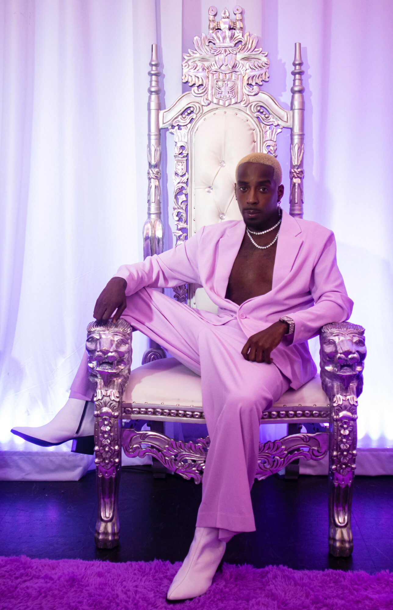 Person in pink suit sitting on ornate throne with white curtain background.