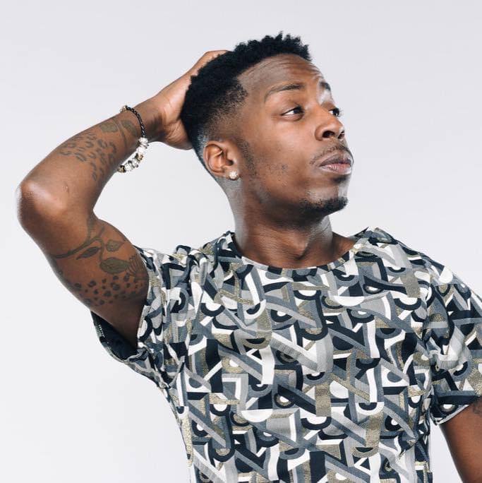 Young man with short hair, wearing a patterned t-shirt, looking to the side with hand on head.