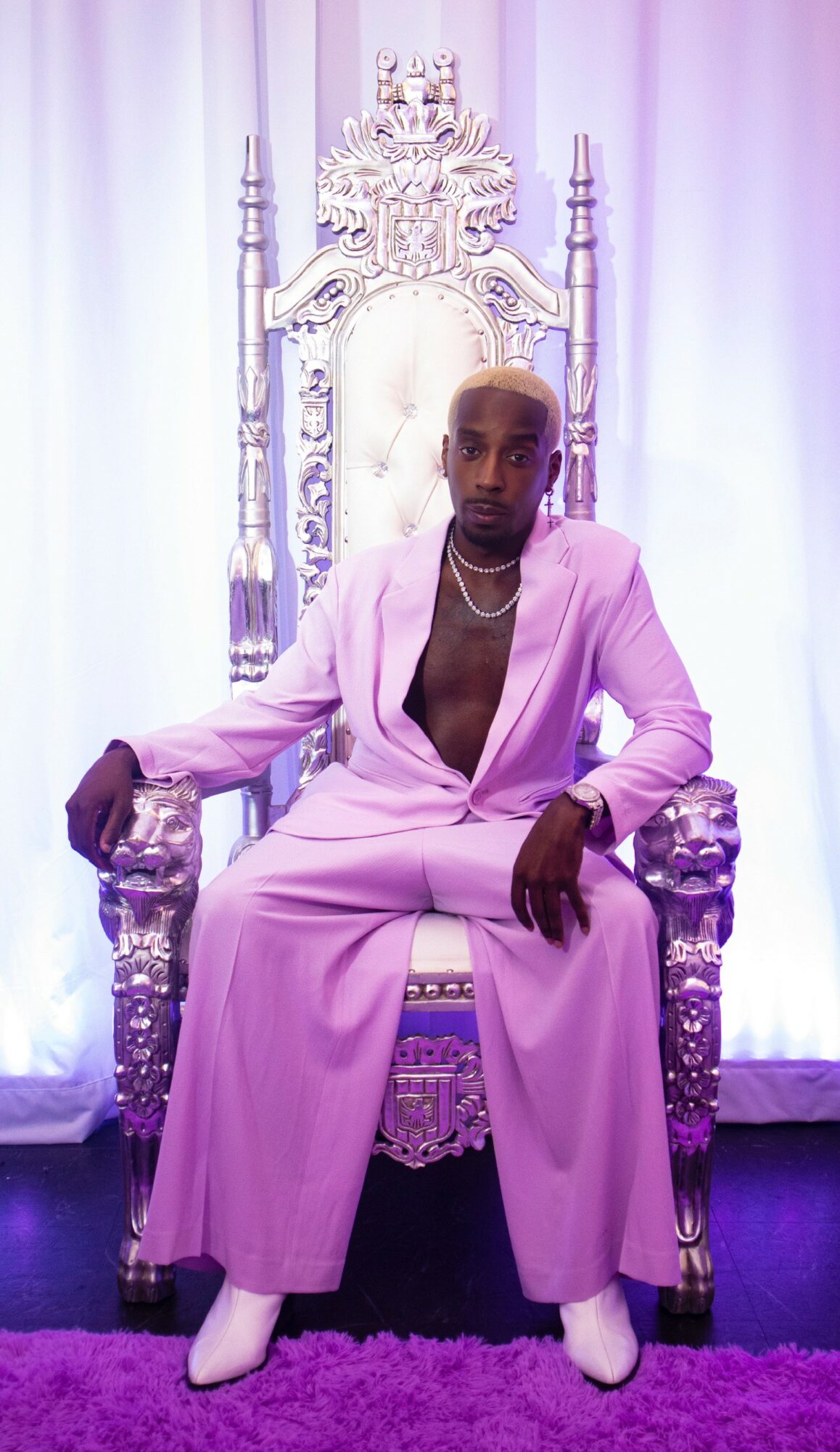 Person seated on ornate throne with pink suit, white shoes, and a necklace, against a backdrop of white and purple curtains.