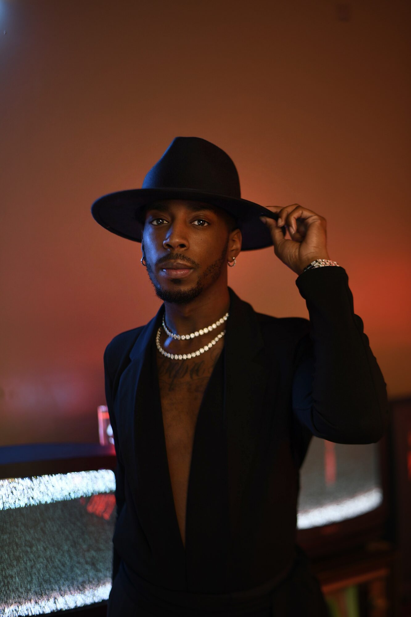 Person wearing a wide-brimmed hat, black outfit, and pearl necklace, holding the hat's brim with one hand, against a dark background.