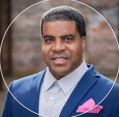 Man in a blue suit with a white shirt and pink pocket square, outdoors with a brick wall background.