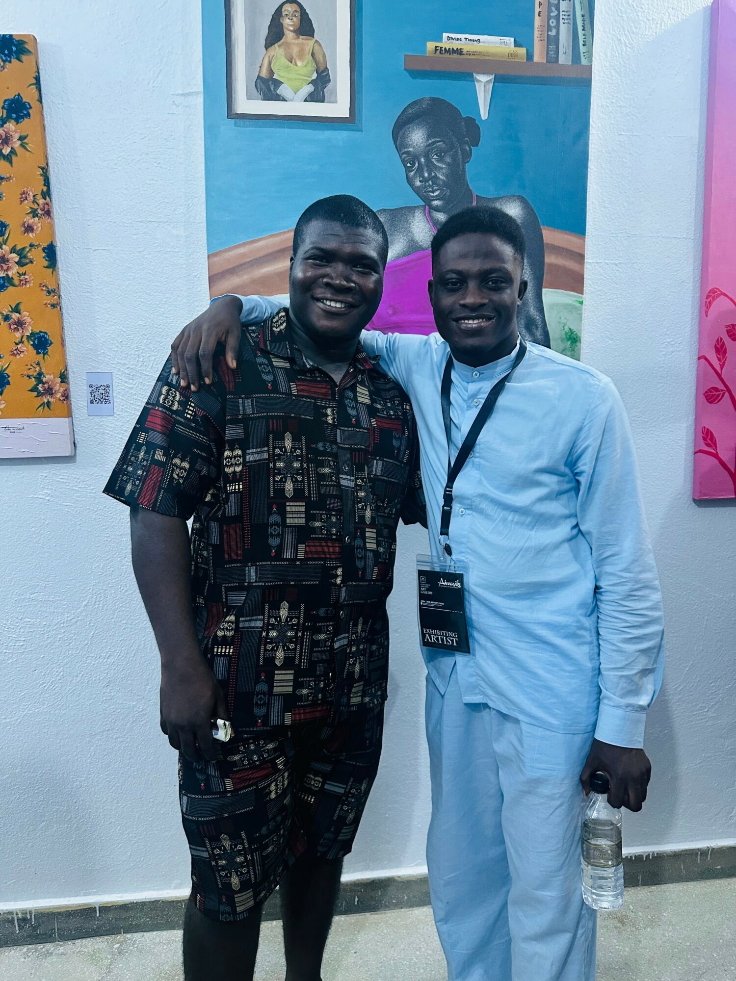 Two men stand together, smiling, one with arm around the other, in an art gallery with colorful paintings behind them.