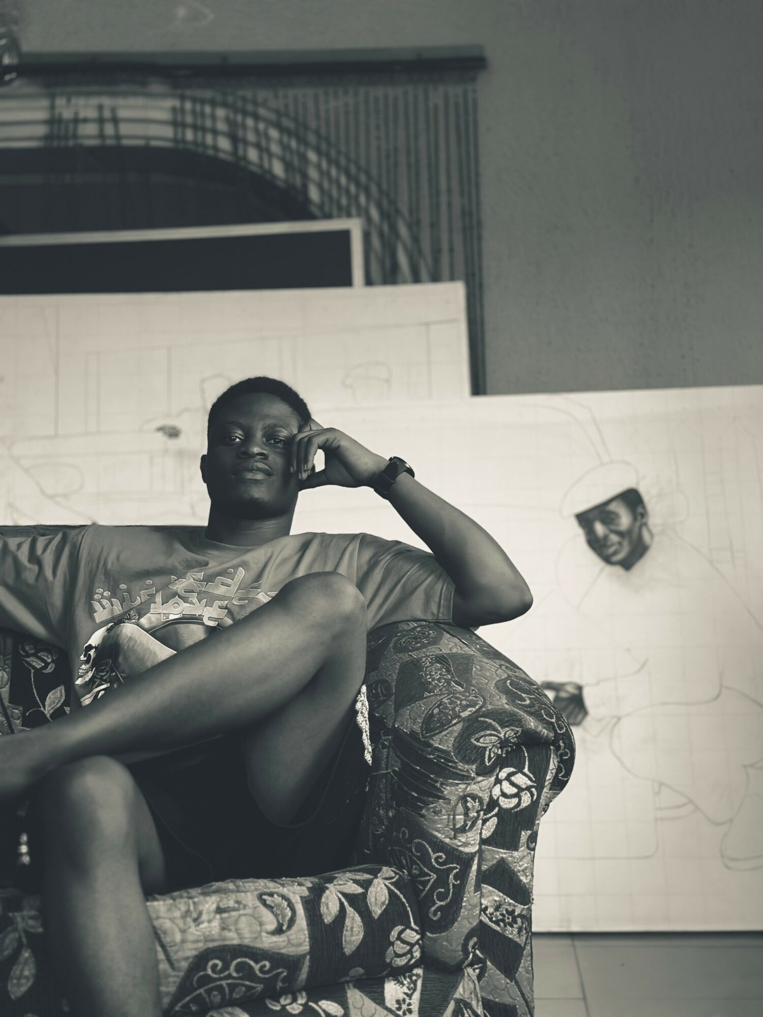 Person sitting on a patterned armchair, legs crossed, with a large drawing or painting behind them.