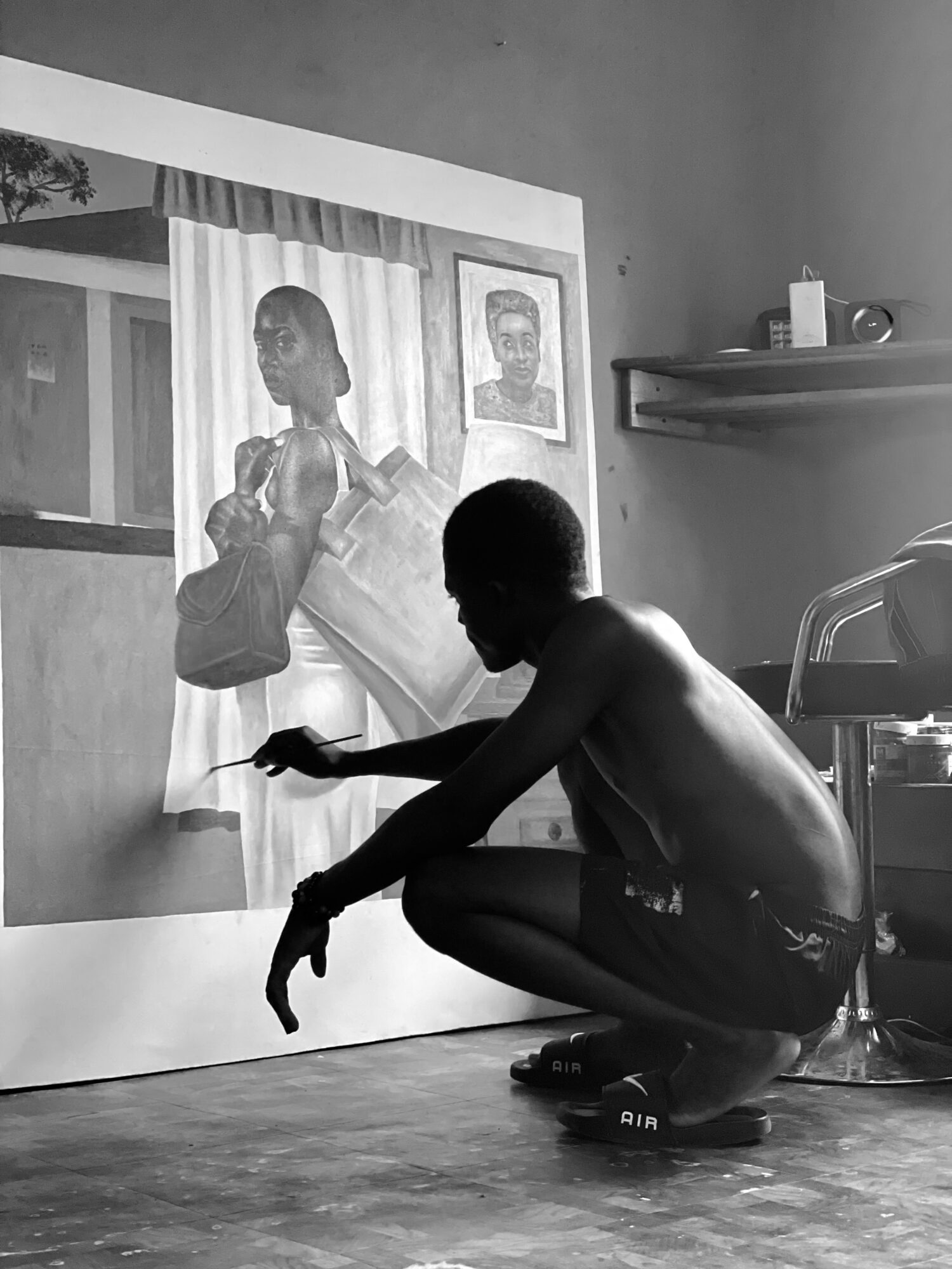 Person crouching in front of a large painting, with a framed picture and shelf on the wall behind.