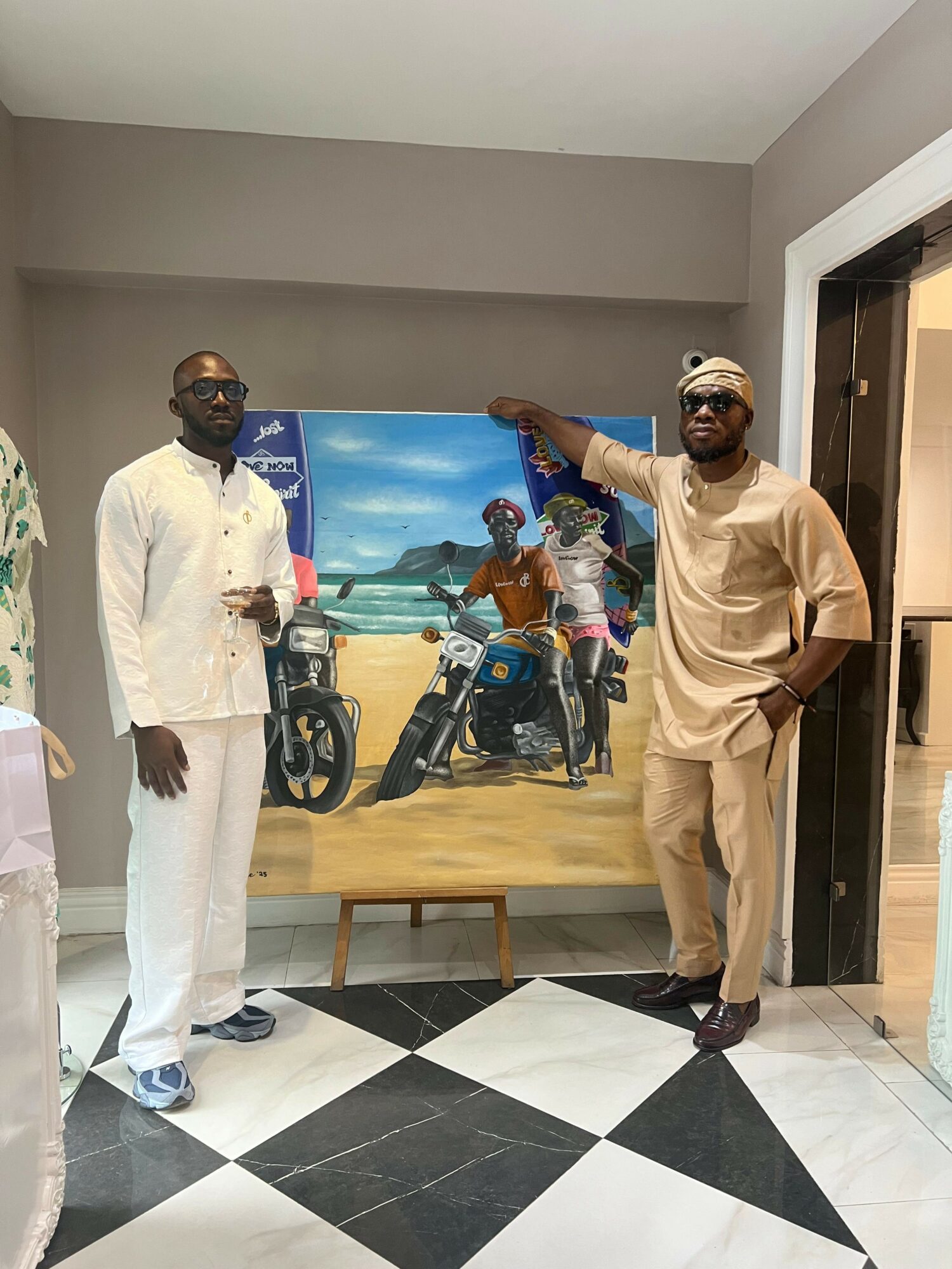 Two men standing indoors in front of a painting of a beach scene with motorcycles and people, one pointing at the artwork.