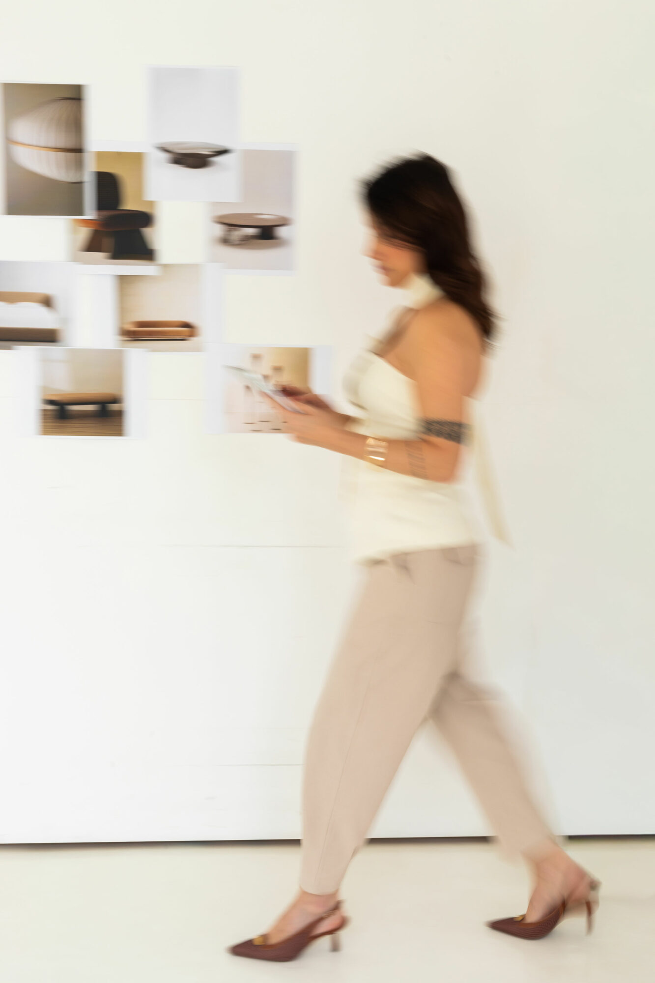 Woman with dark hair in beige pants and heels reads a book in front of a white wall with shelves.