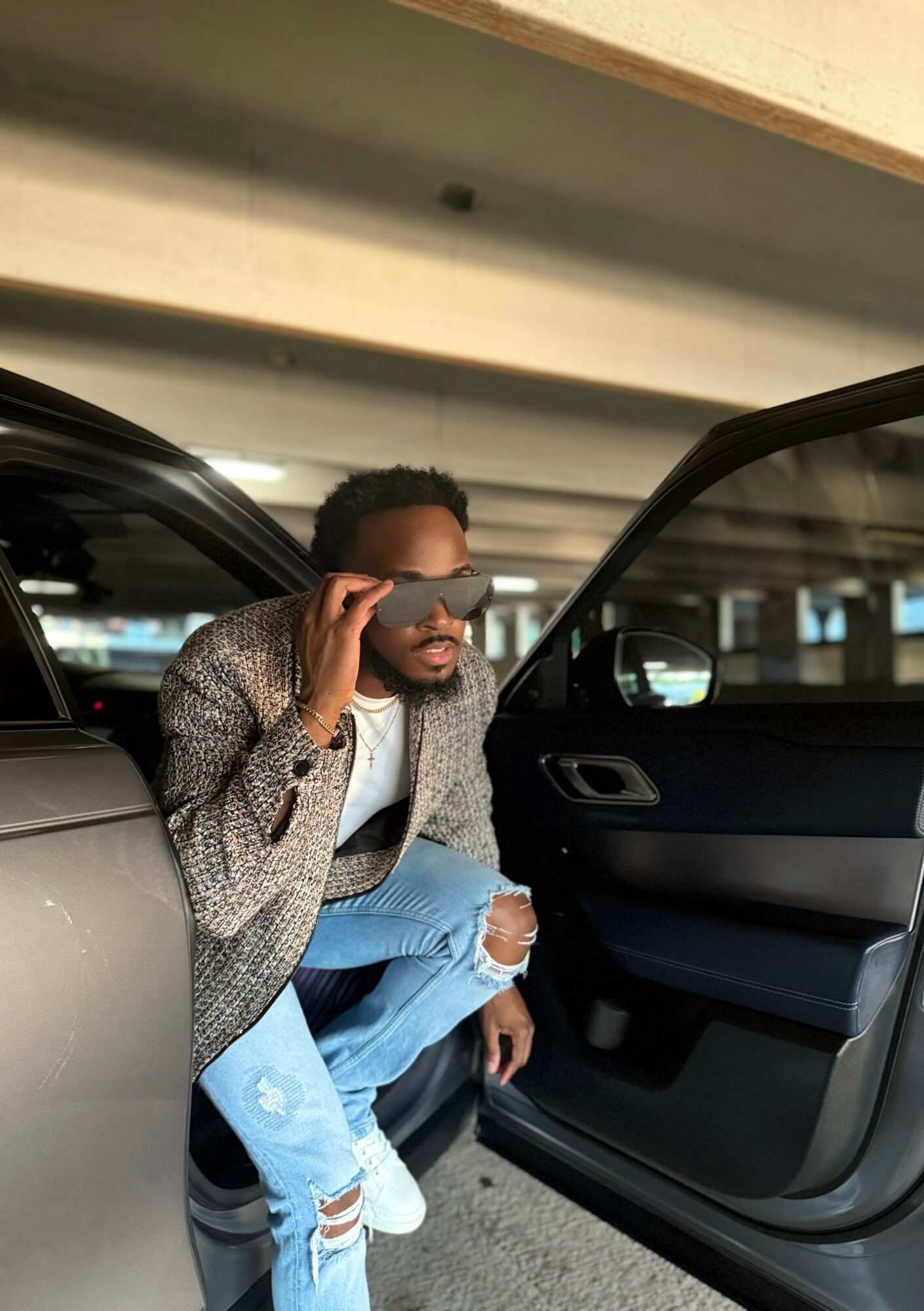 Person sitting in car with door open, looking into distance, wearing glasses, patterned jacket, ripped jeans, in parking garage.
