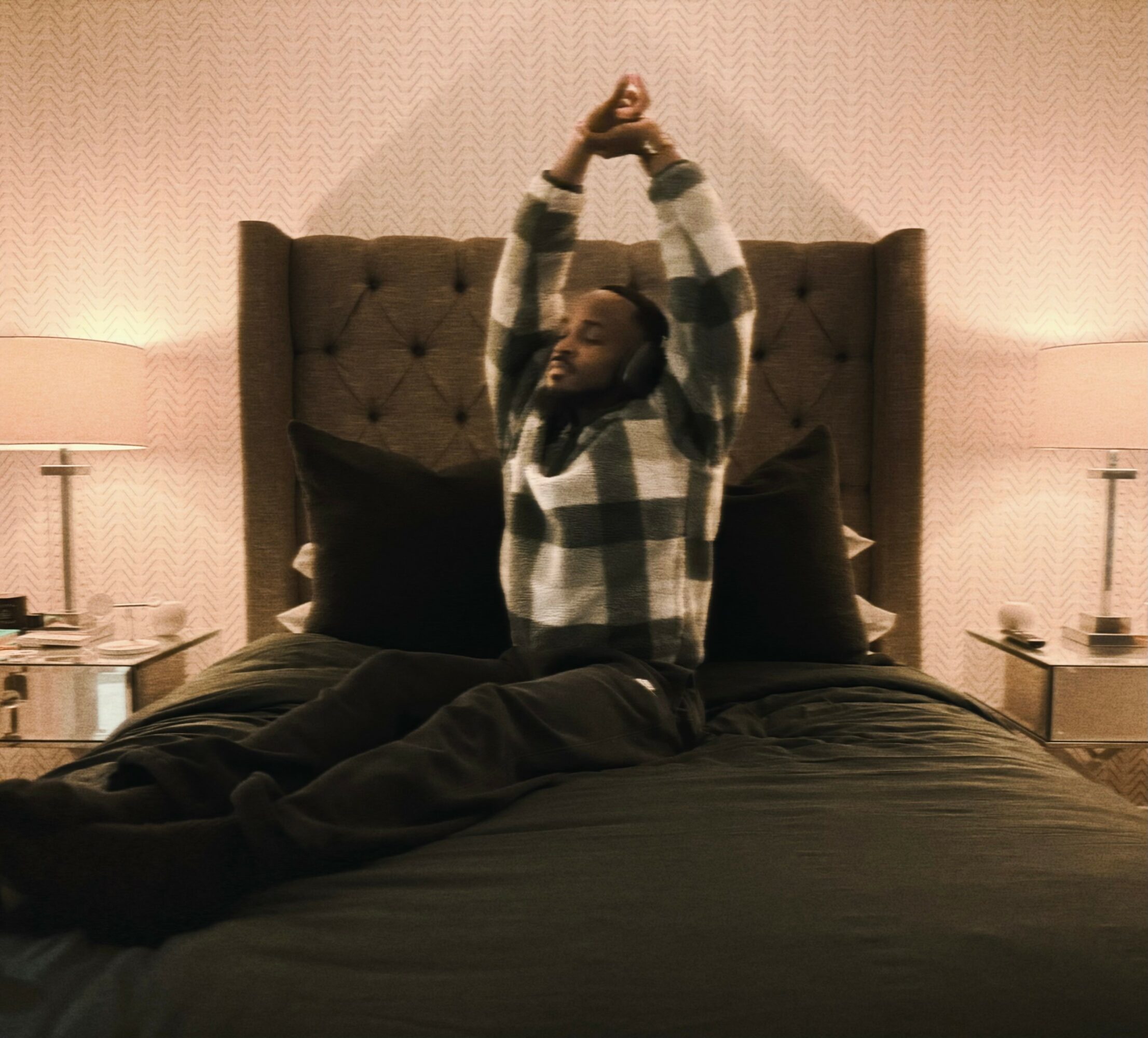 Person sitting on bed with arms raised, in a bedroom with two lamps and nightstands, head tilted back.