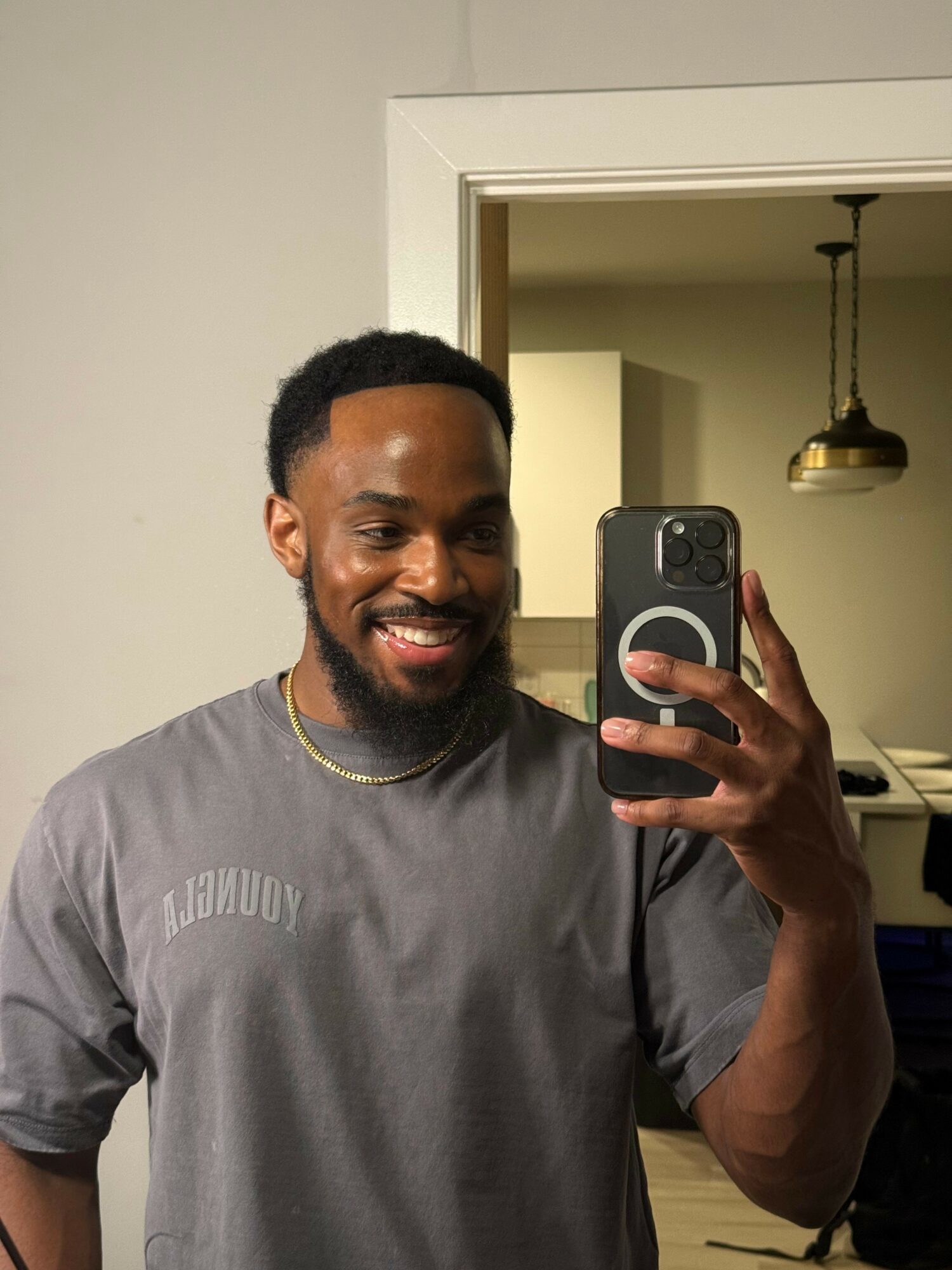 Smiling man taking a selfie in a mirror, wearing a gray shirt, in a room with a doorway and hanging light.