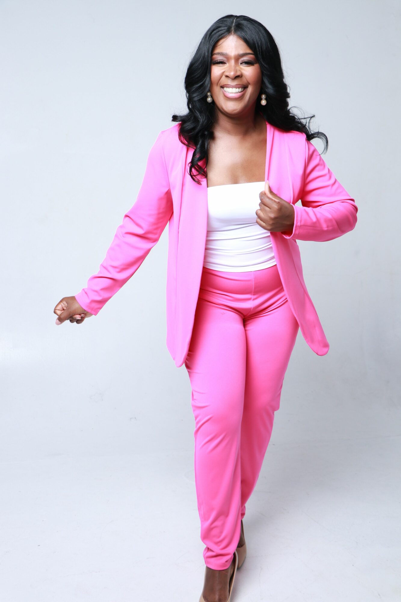 Woman in pink suit and white top smiling, walking forward against a plain background.