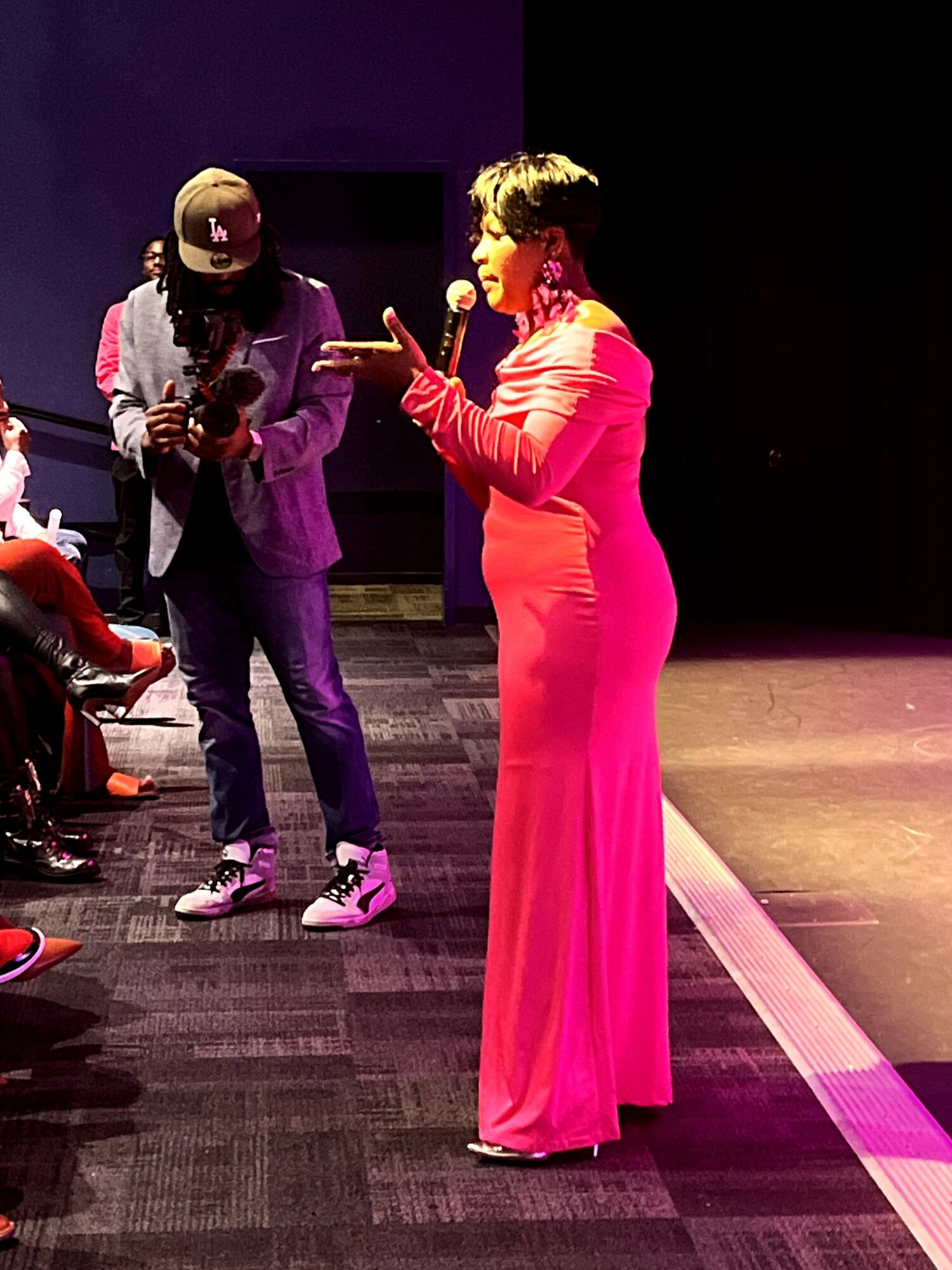 Woman in pink dress speaking into a microphone on stage, with a person in casual clothing nearby.