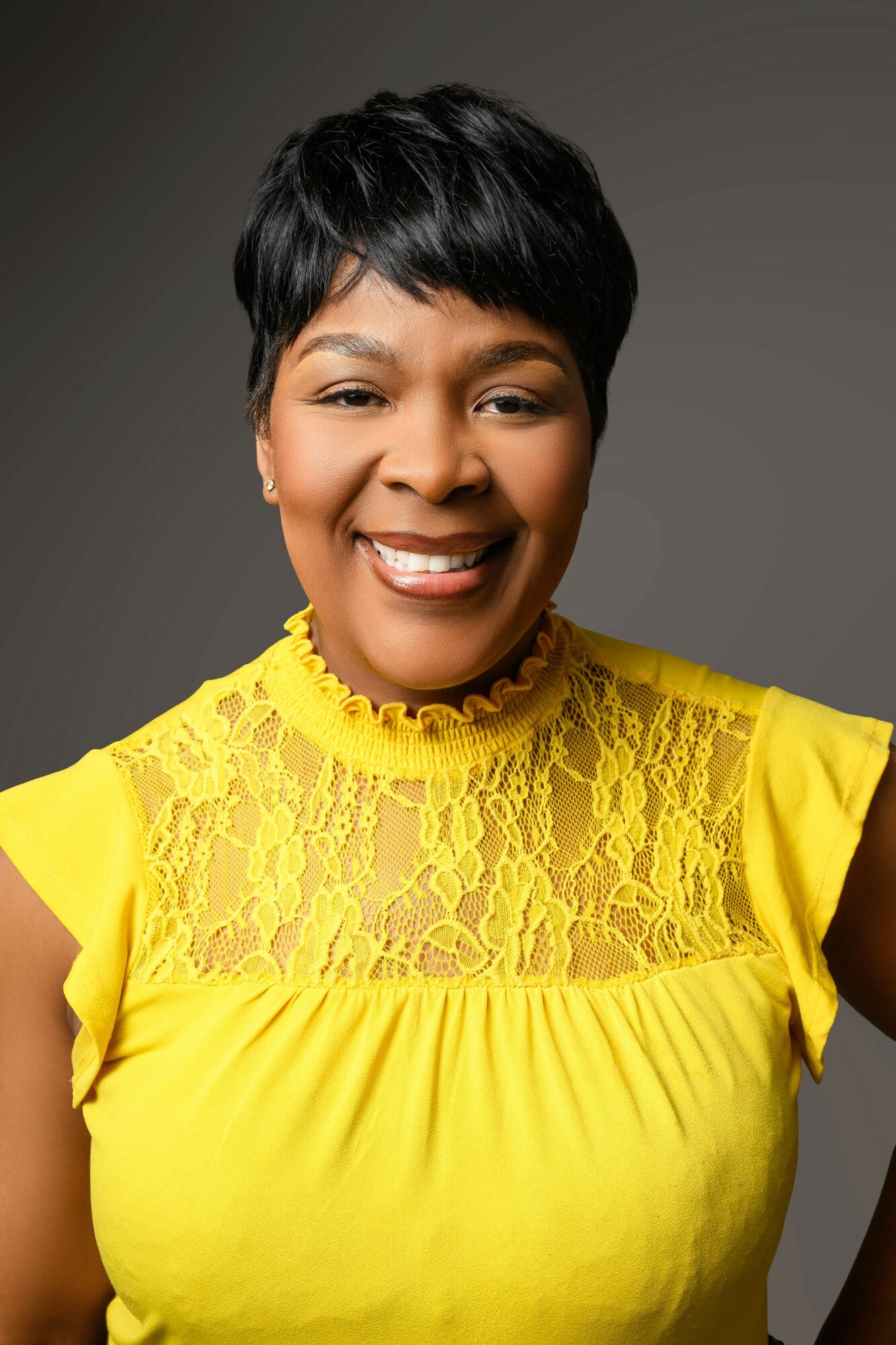 Smiling woman with short black hair wearing a yellow lace top against a gray background.