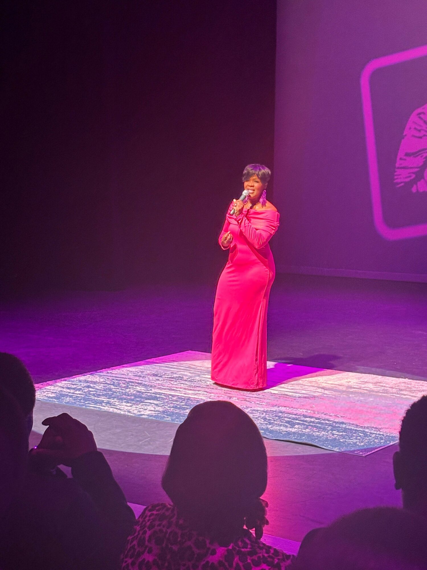 Woman in pink dress standing on stage with microphone, audience in foreground, purple background with screen projection.