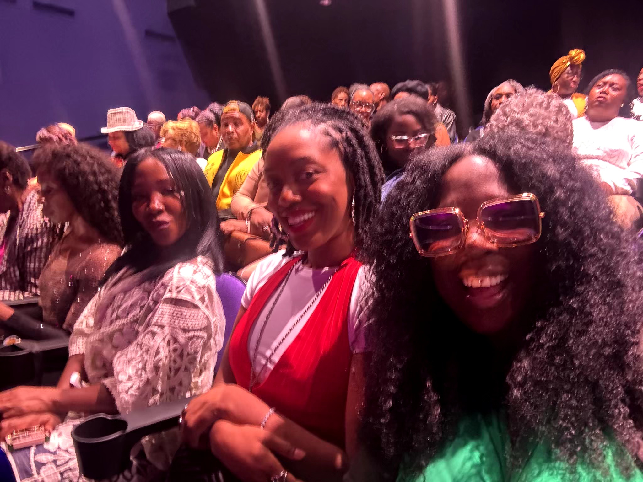 Group of smiling women sitting together at an indoor event, some wearing glasses and colorful clothing.