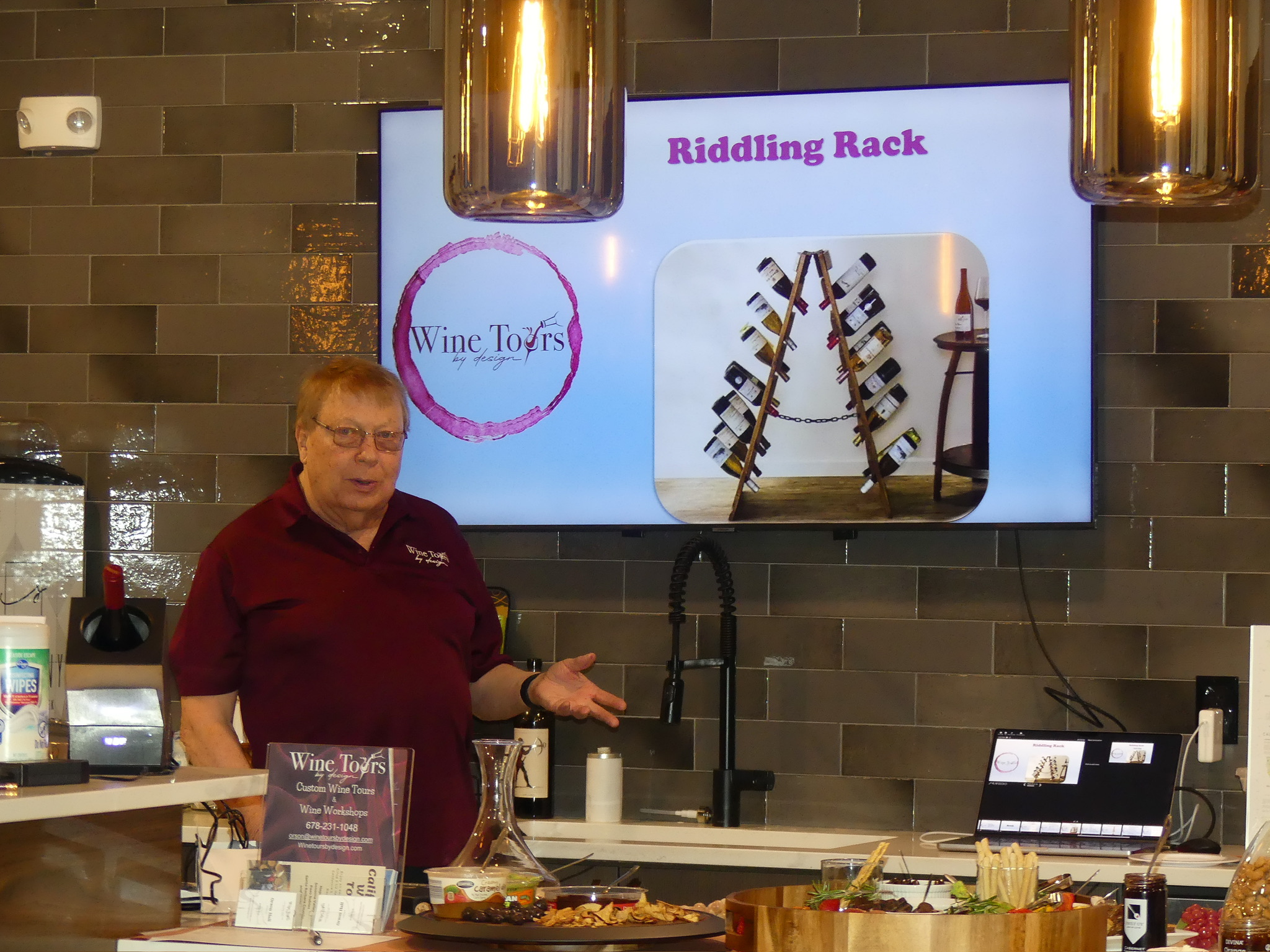 Woman standing behind a counter with a screen displaying a Riddling Rack and Wine Tours logo in a store or tasting room.