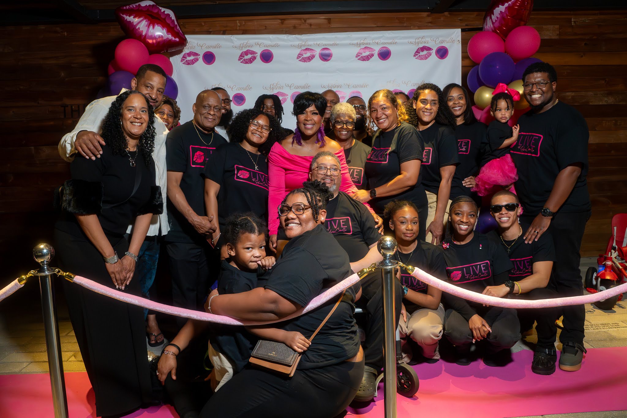 Group of women and children celebrating with pink and red balloons and decorations, smiling together indoors.