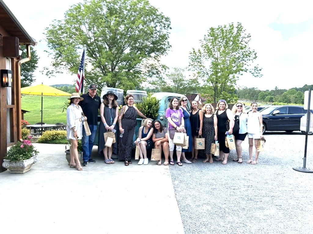 Group of people standing outdoors in front of trees and parked cars, some holding umbrellas, on a sunny day.