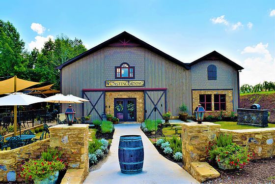 Barn-style building with a pathway leading to entrance, surrounded by plants and outdoor seating, under a blue sky.