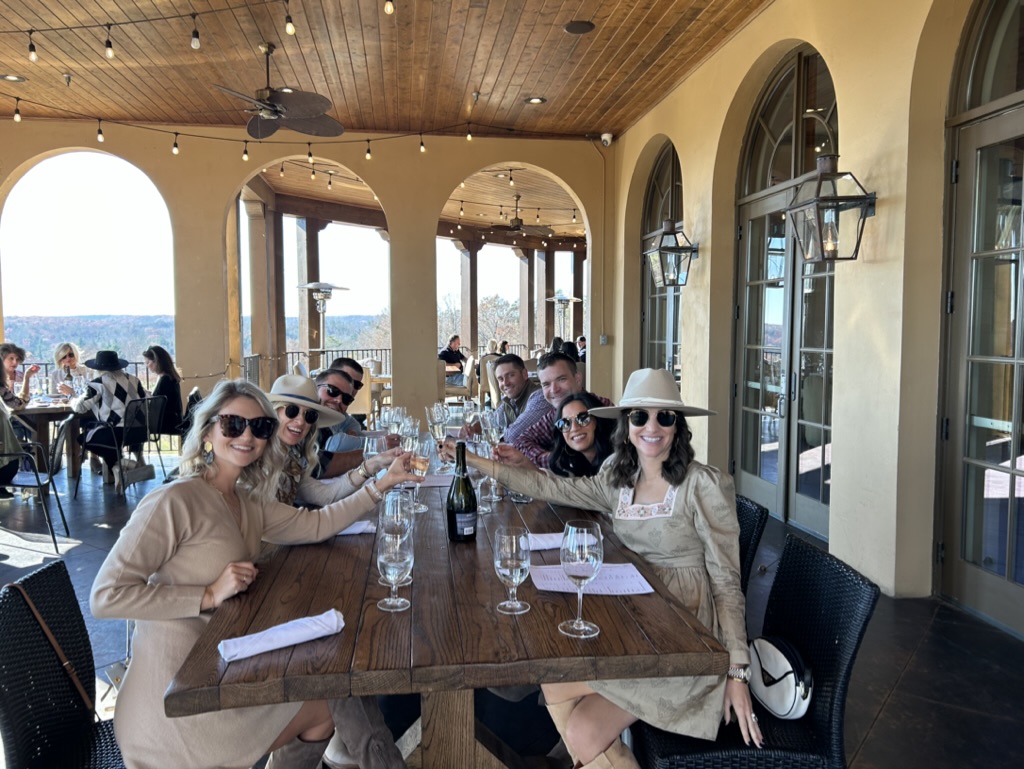 Group of people sitting at a long table on a patio with large windows and a view of the ocean.