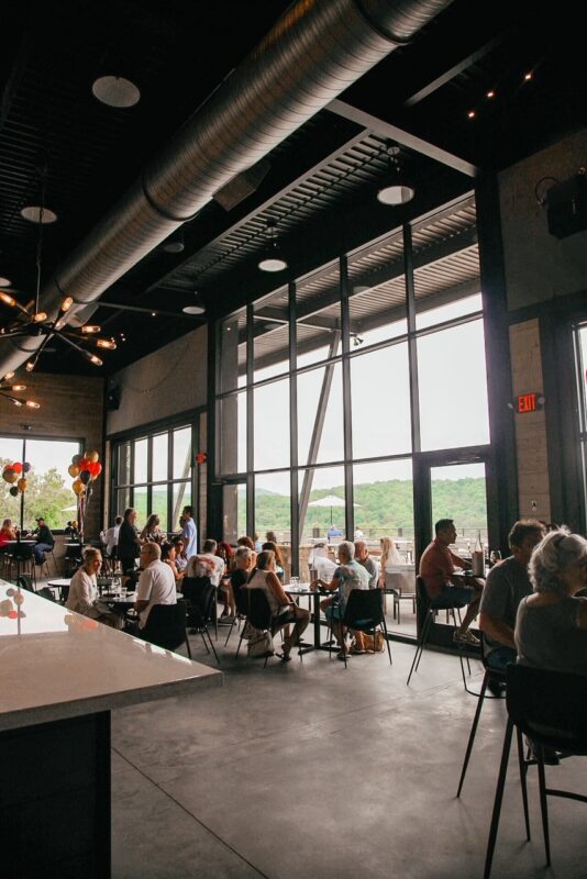 People sit at tables near large windows in a modern restaurant or cafe with industrial ceiling design.
