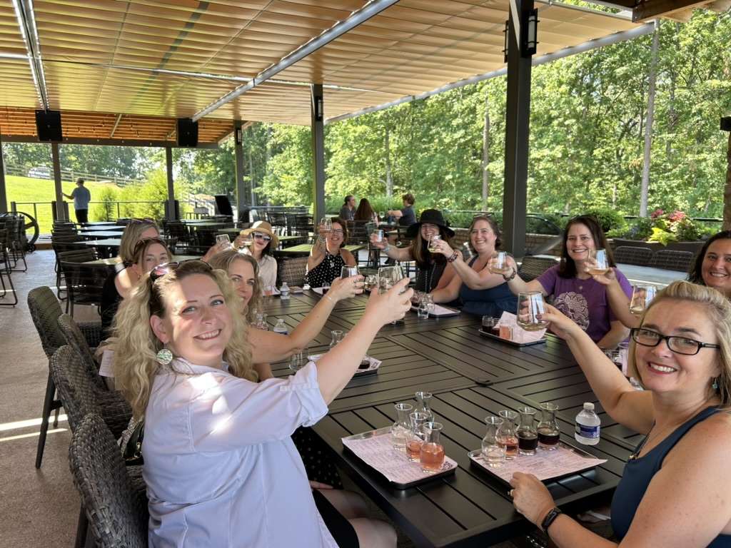 Group of women sitting at a long outdoor table, smiling and raising glasses in a toast, with green trees in background.