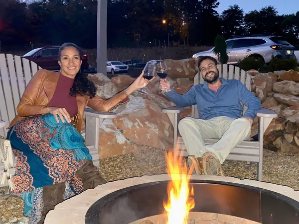 Two people sitting outdoors near a fire pit, clinking glasses of red wine, smiling, with cars and trees in background.