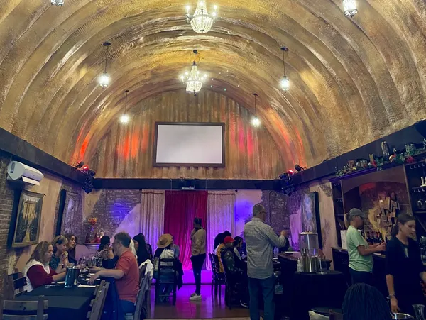 People gather in a decorated venue with a curved ceiling, chandeliers, and a large screen, some seated and others standing.