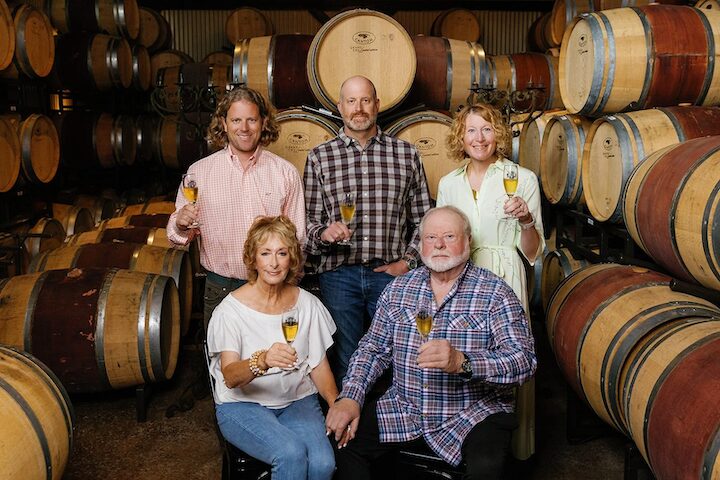 Five people in a wine cellar holding glasses of wine, surrounded by stacked wine barrels.