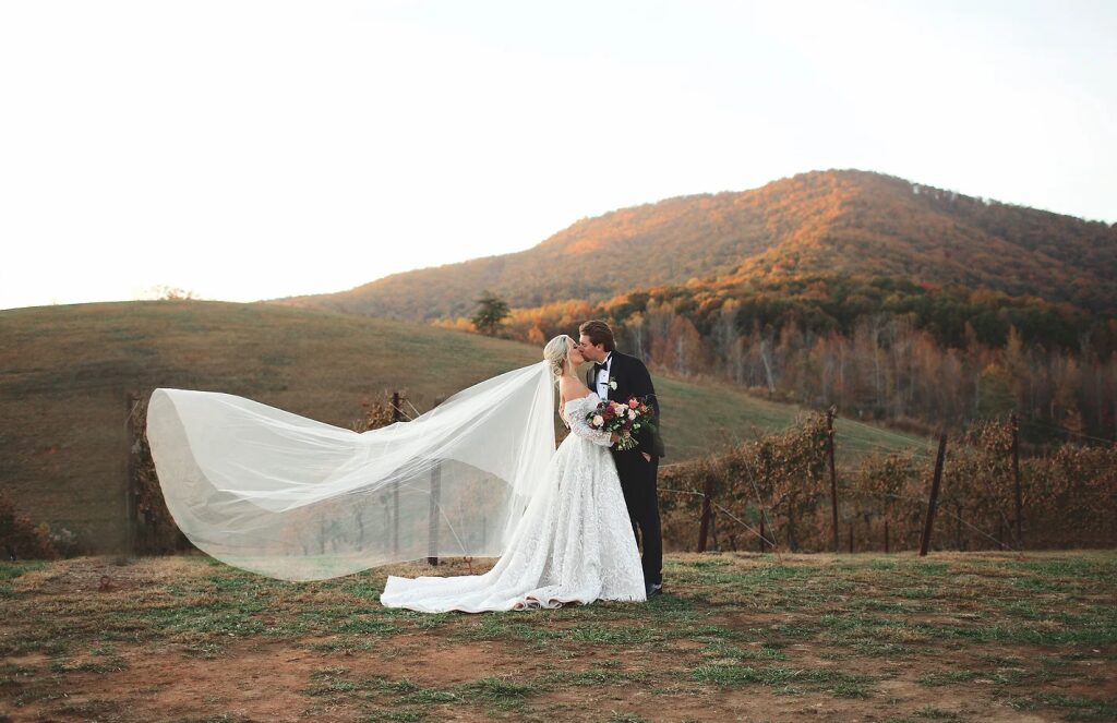 Bride and groom standing outdoors with a scenic hilly landscape in the background, bride holding a bouquet, bride's veil flowing.