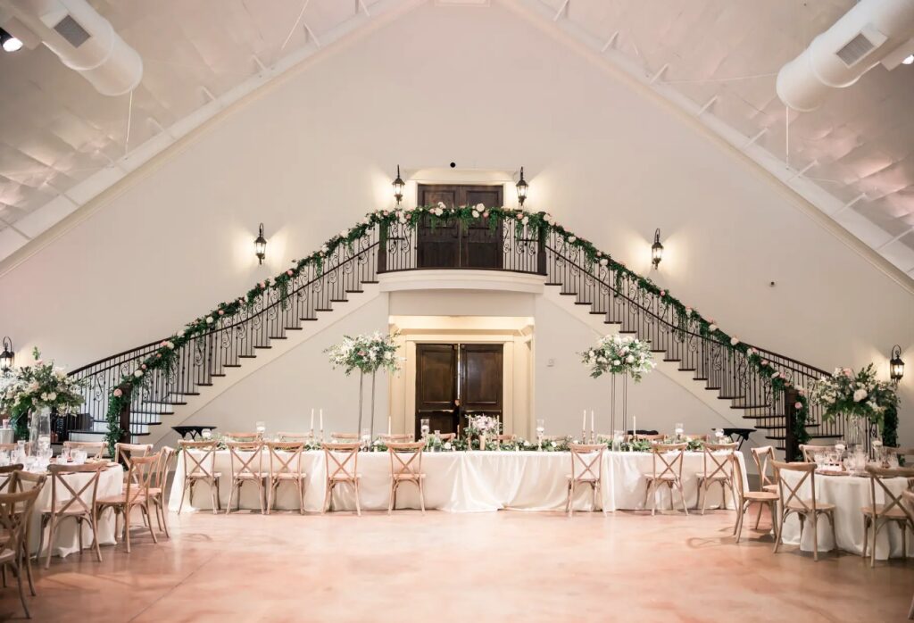 Elegant banquet hall with a high ceiling, staircase, and decorated tables with chairs, floral arrangements, and lighting fixtures.