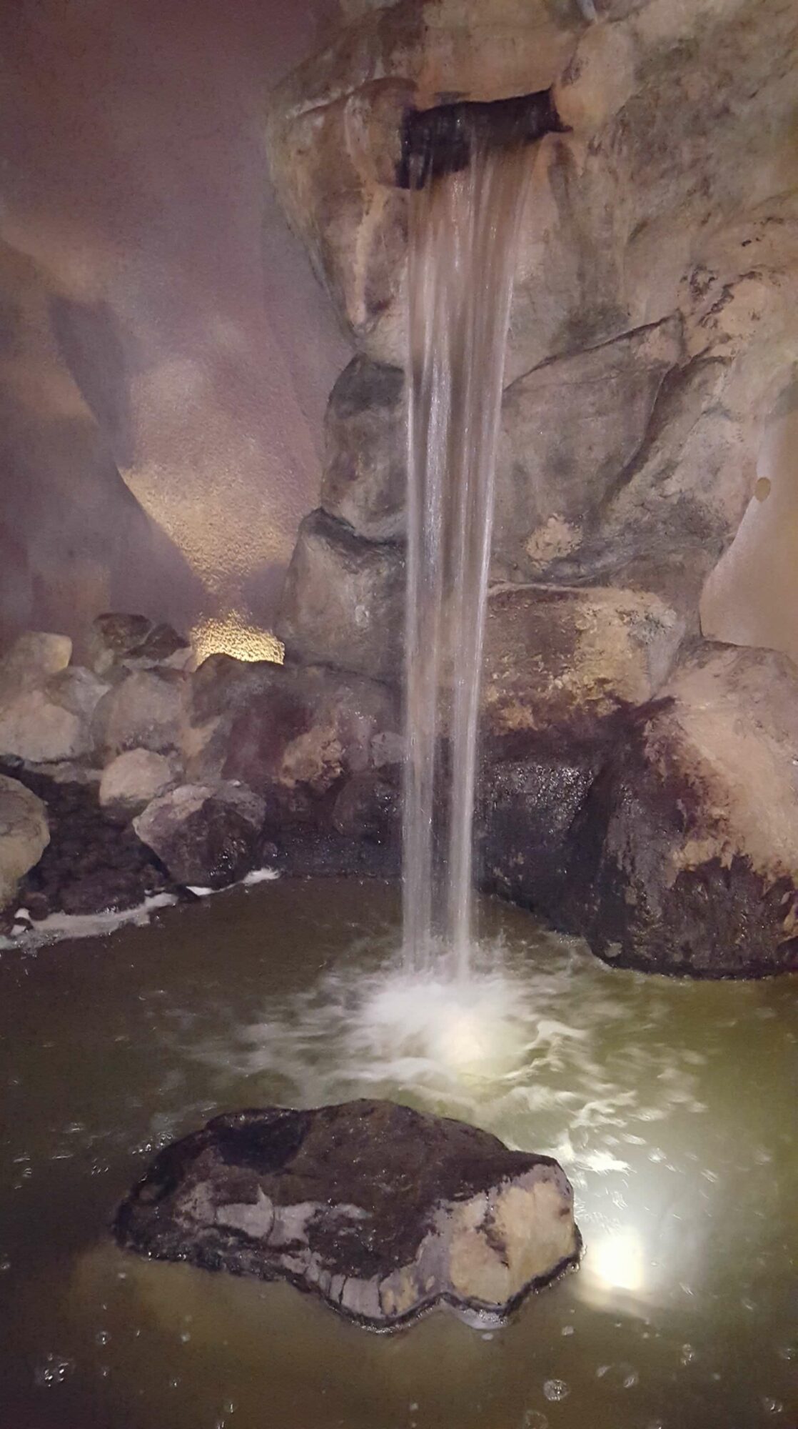 Water flows from a narrow opening between rocks into a pool, with mist and rocks visible around the water source.