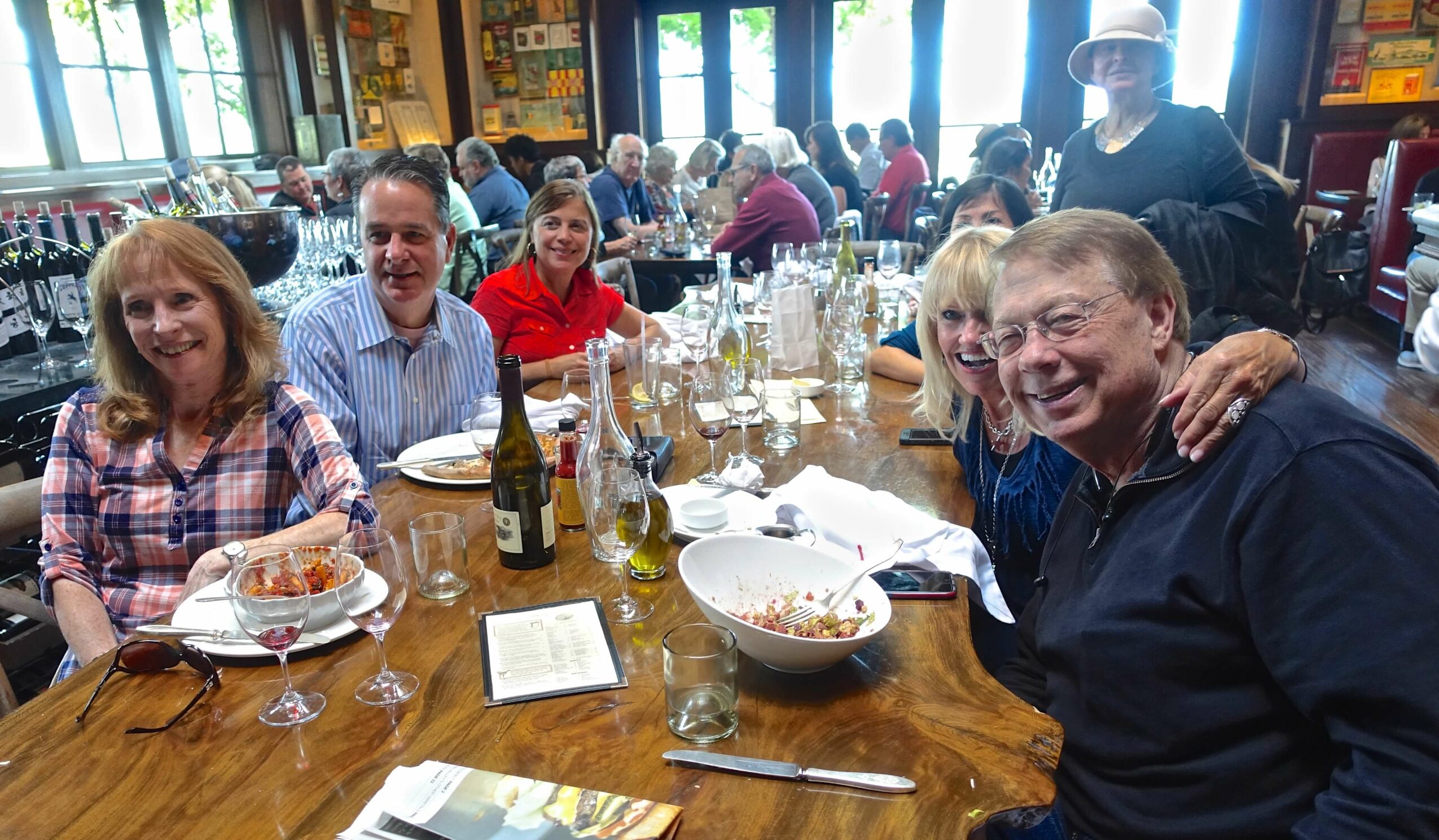 Group of six people sitting around a wooden table with food and drinks, smiling and enjoying a meal in a lively restaurant.
