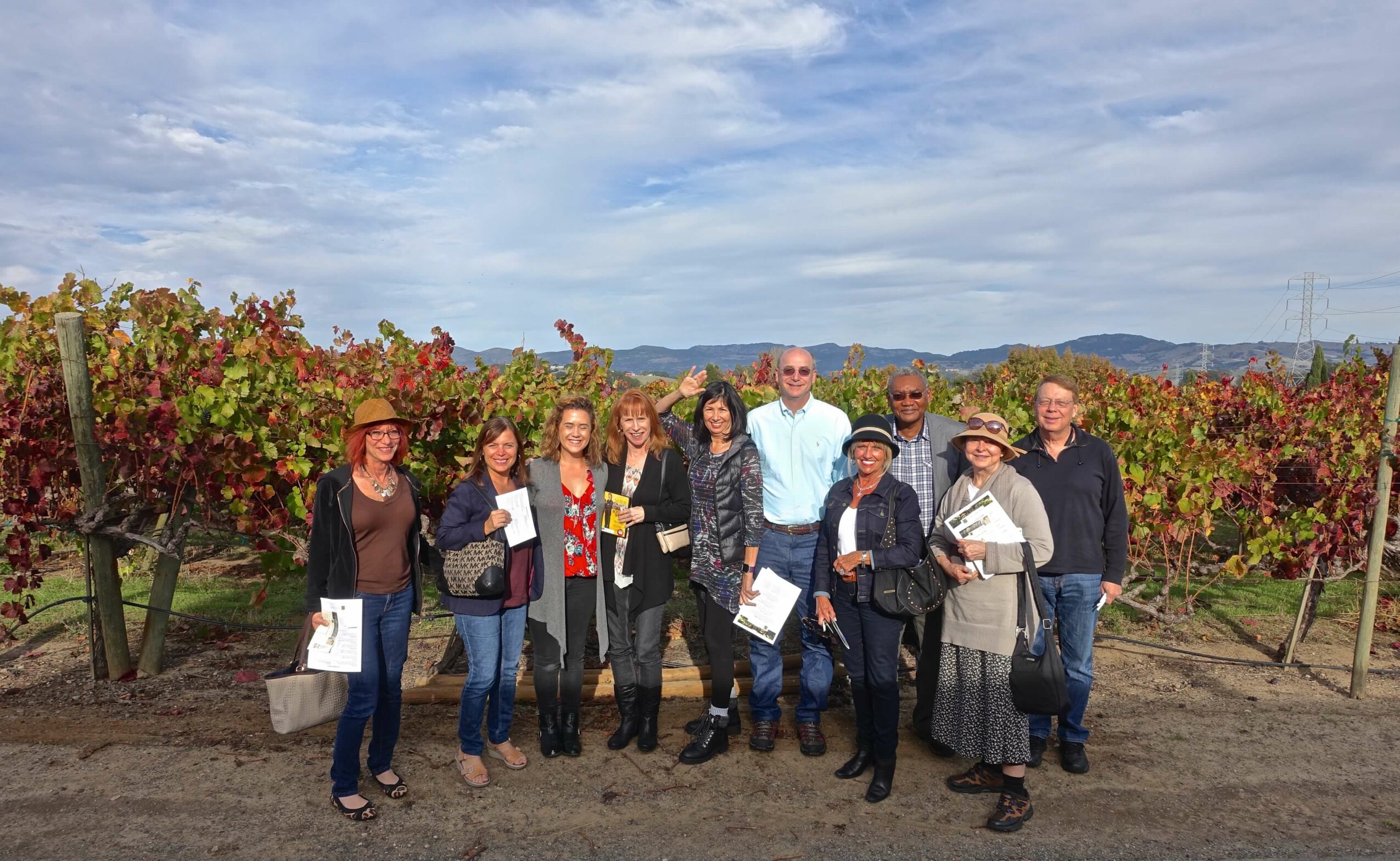 Group of ten people standing outdoors in a vineyard with grapevines and hills in the background.