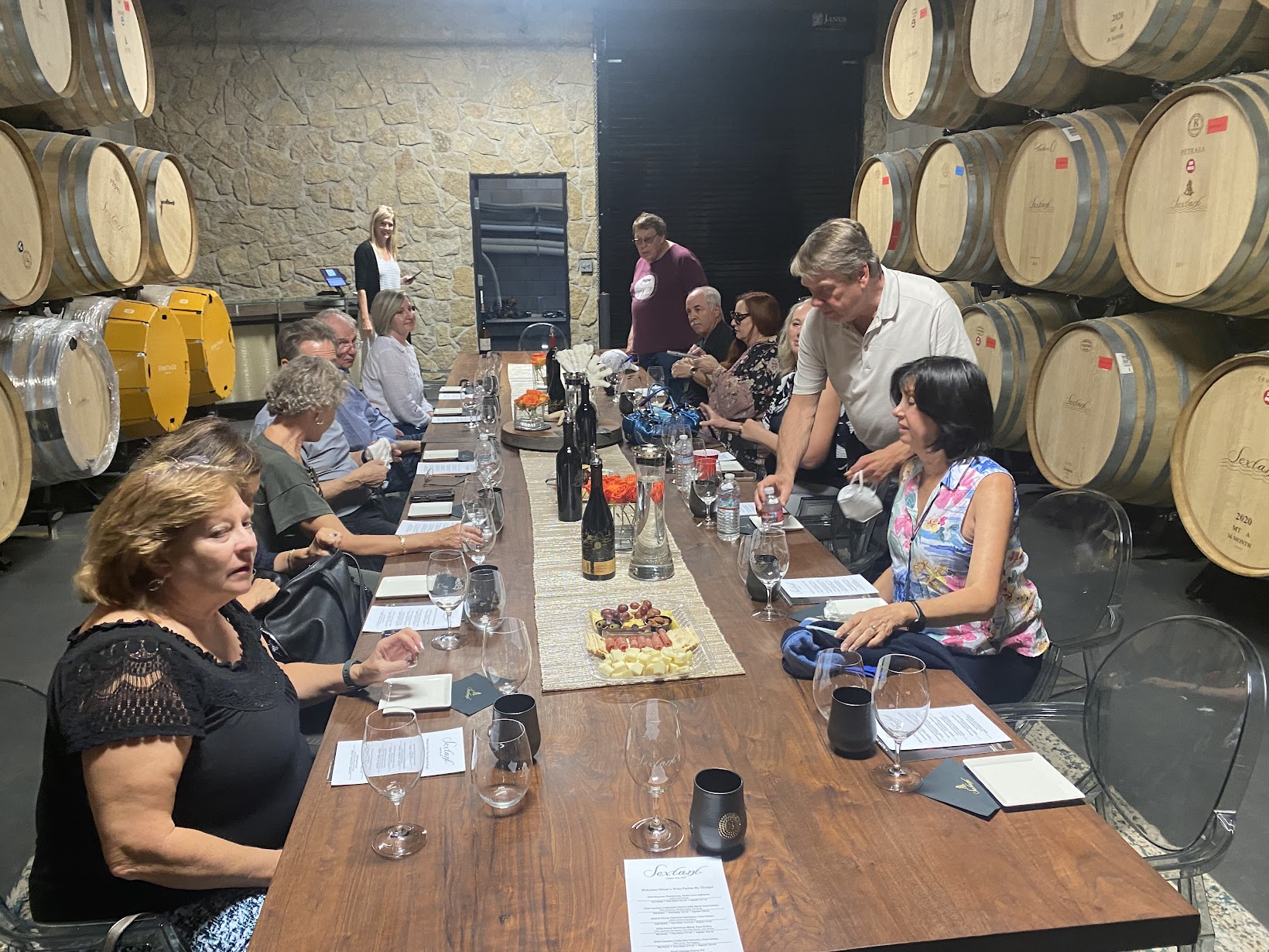 People gathered around a long table in a wine cellar with barrels on walls, tasting and discussing wine.