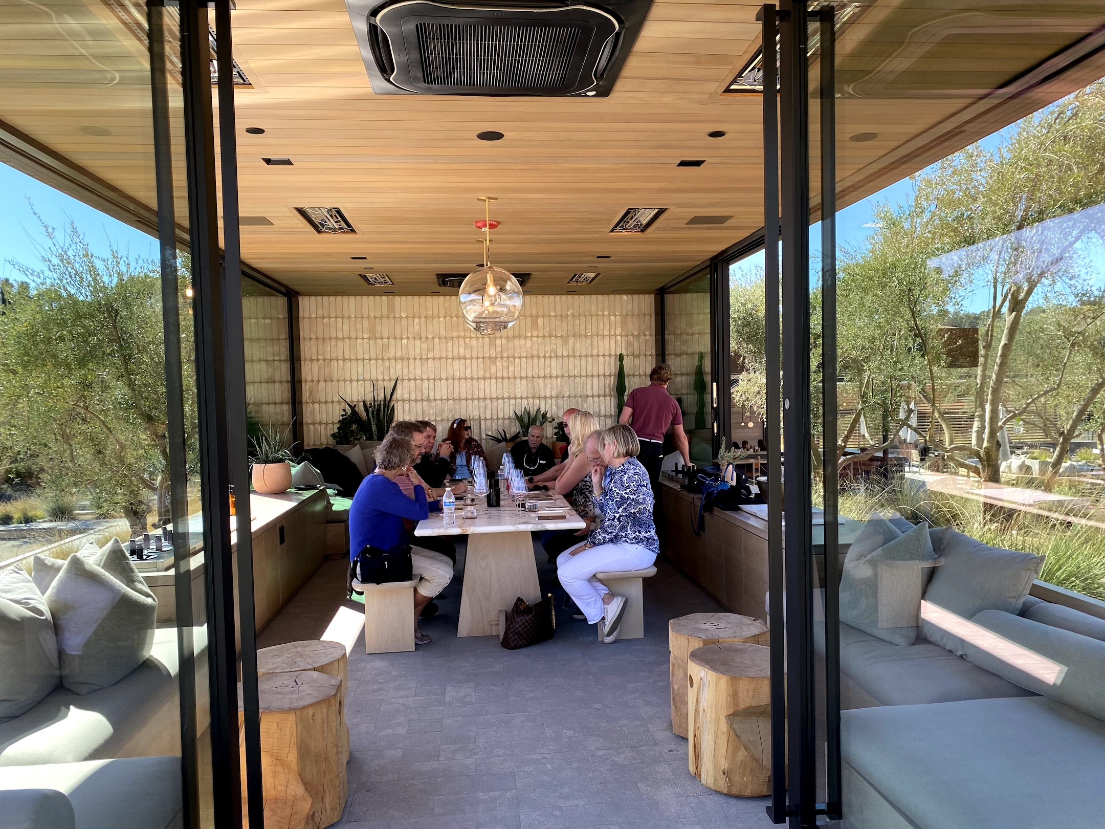 People sit around a table in a room with large windows, trees visible outside, and wooden ceiling with a chandelier.